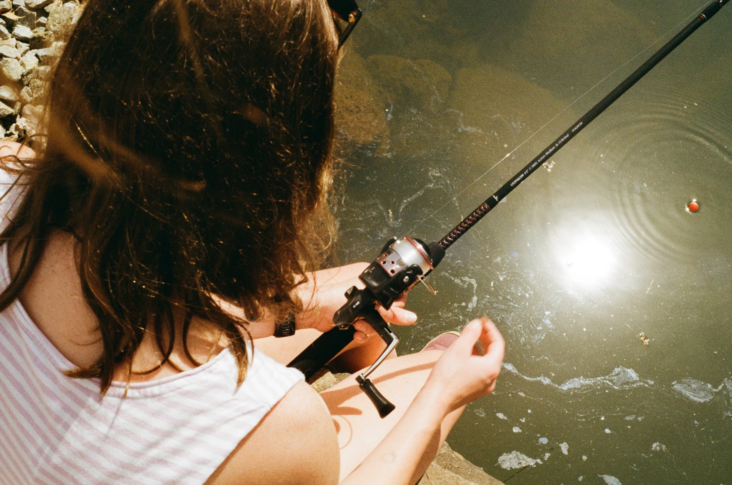 A person with wavy brown hair, wearing glasses and a striped sleeveless top, is fishing with a spinning rod and reel at a body of water near a rocky shore.