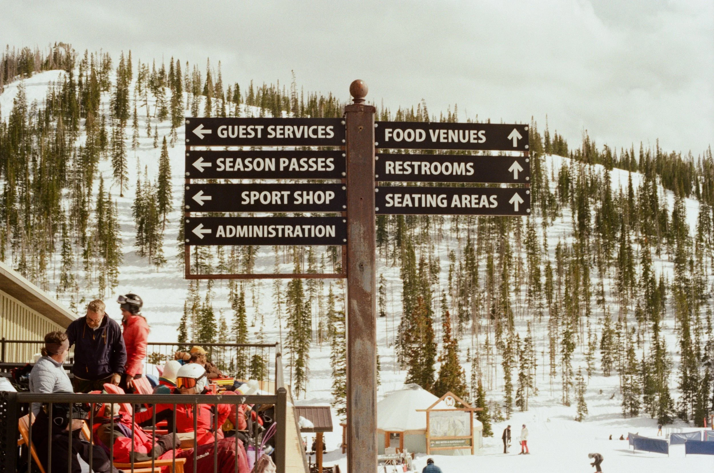 Signpost at a ski resort with directions to guest services, food venues, season passes, restrooms, sport shop, seating areas, and administration. People in ski gear and snow-covered trees in the background.