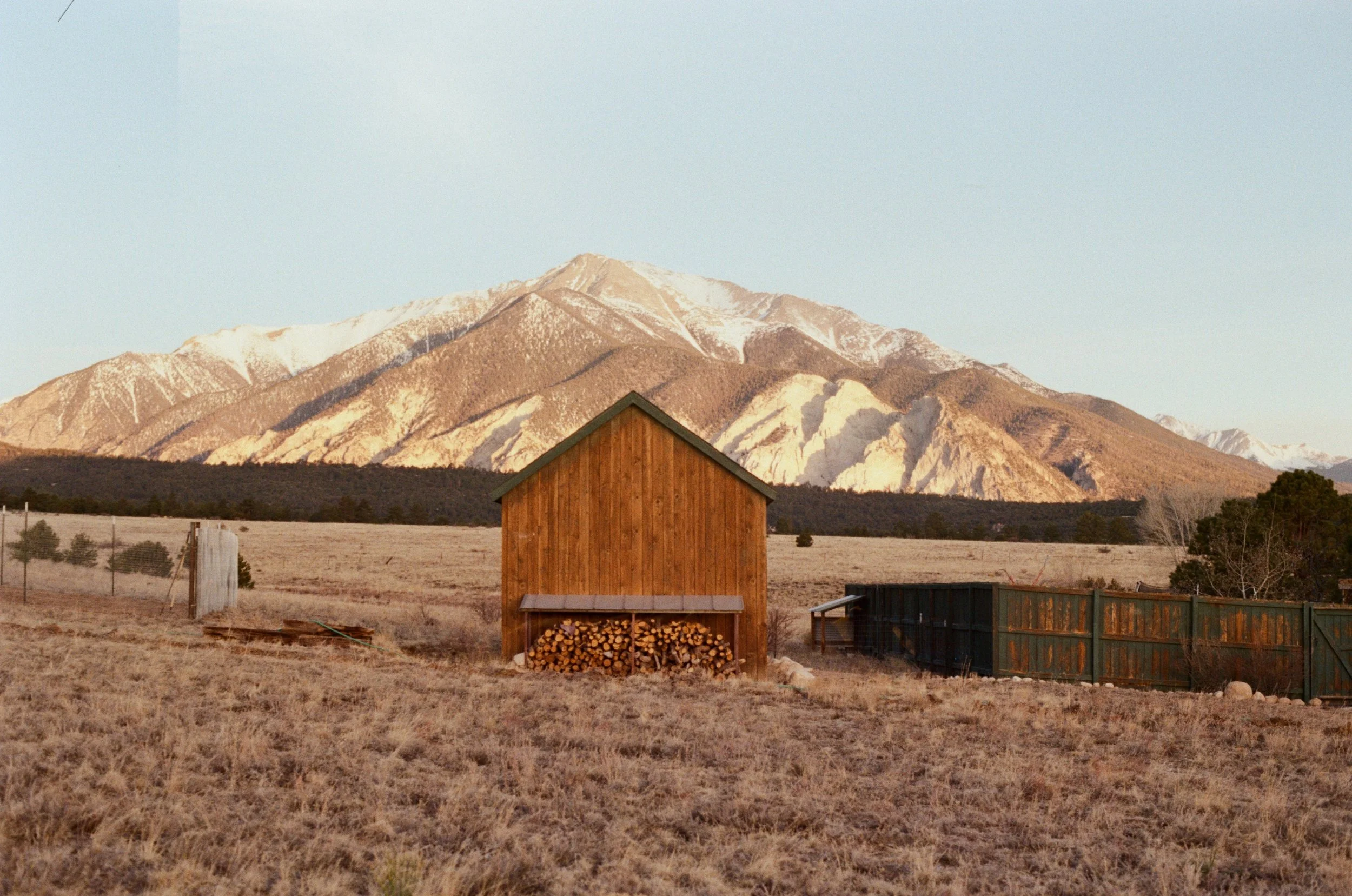 A small wooden shed with a stack of firewood underneath a covered porch, set in a rural field with a mountain range in the background.