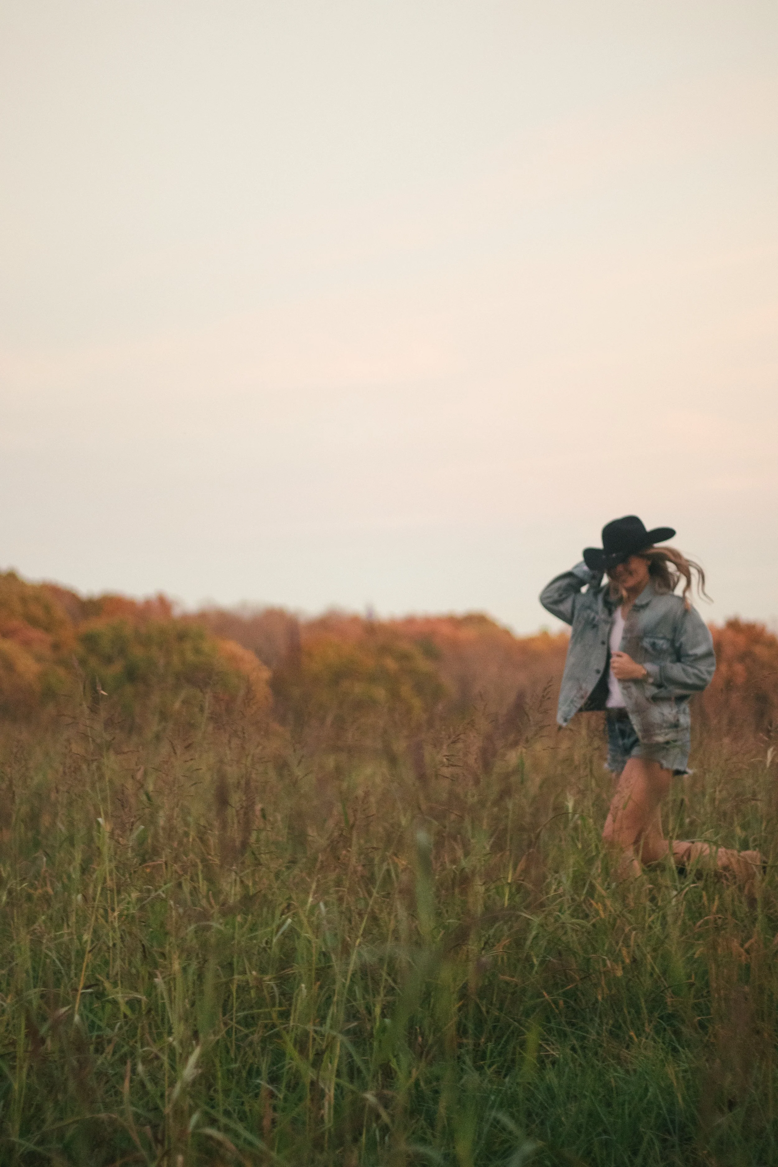 A woman wearing a black hat and denim jacket walking through a grassy field with trees in the background during sunset or sunrise.