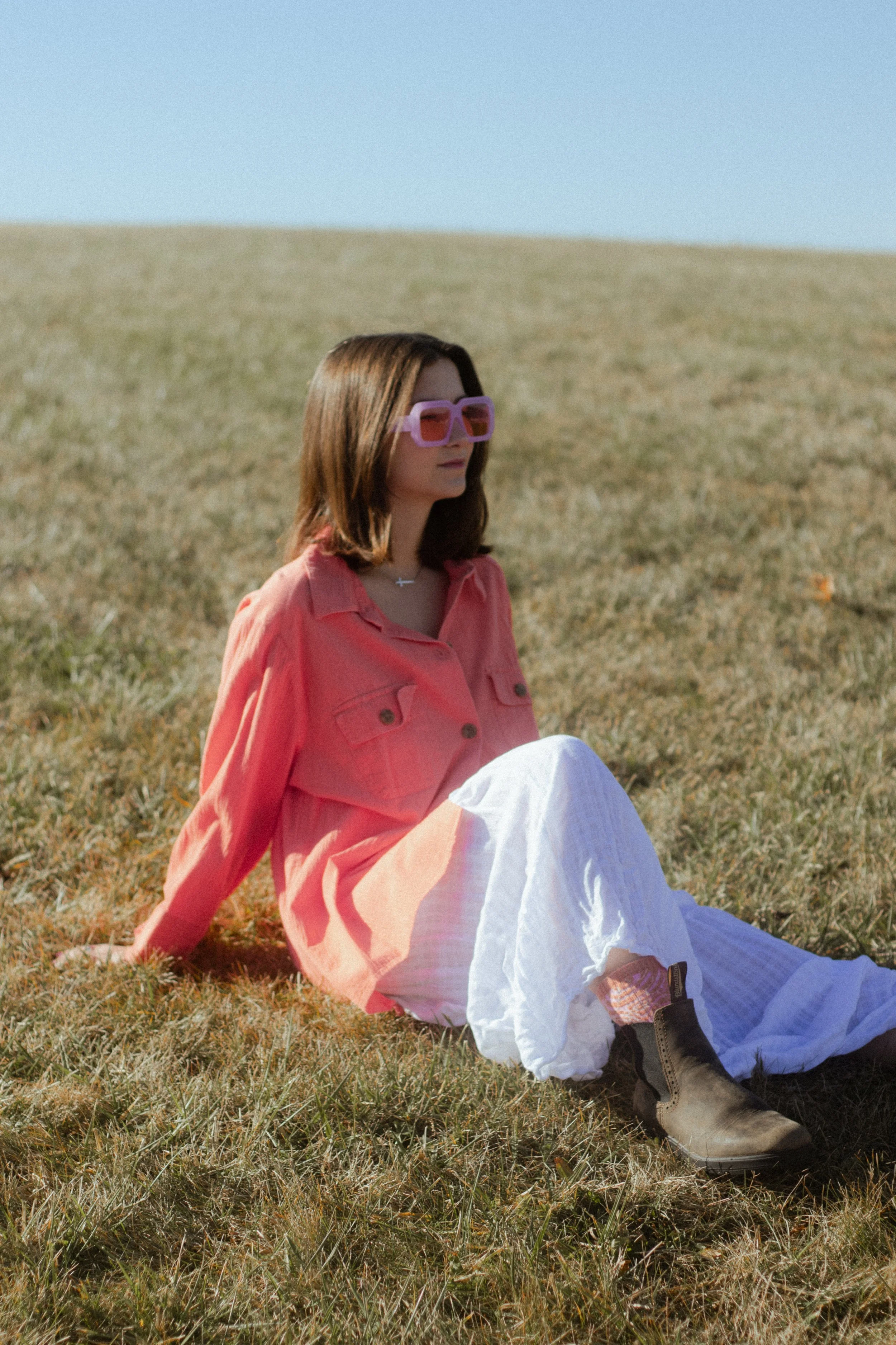 A woman with shoulder-length brown hair wearing pink sunglasses, a coral pink jacket, and white pants, sitting on grass in an open field with a clear blue sky.