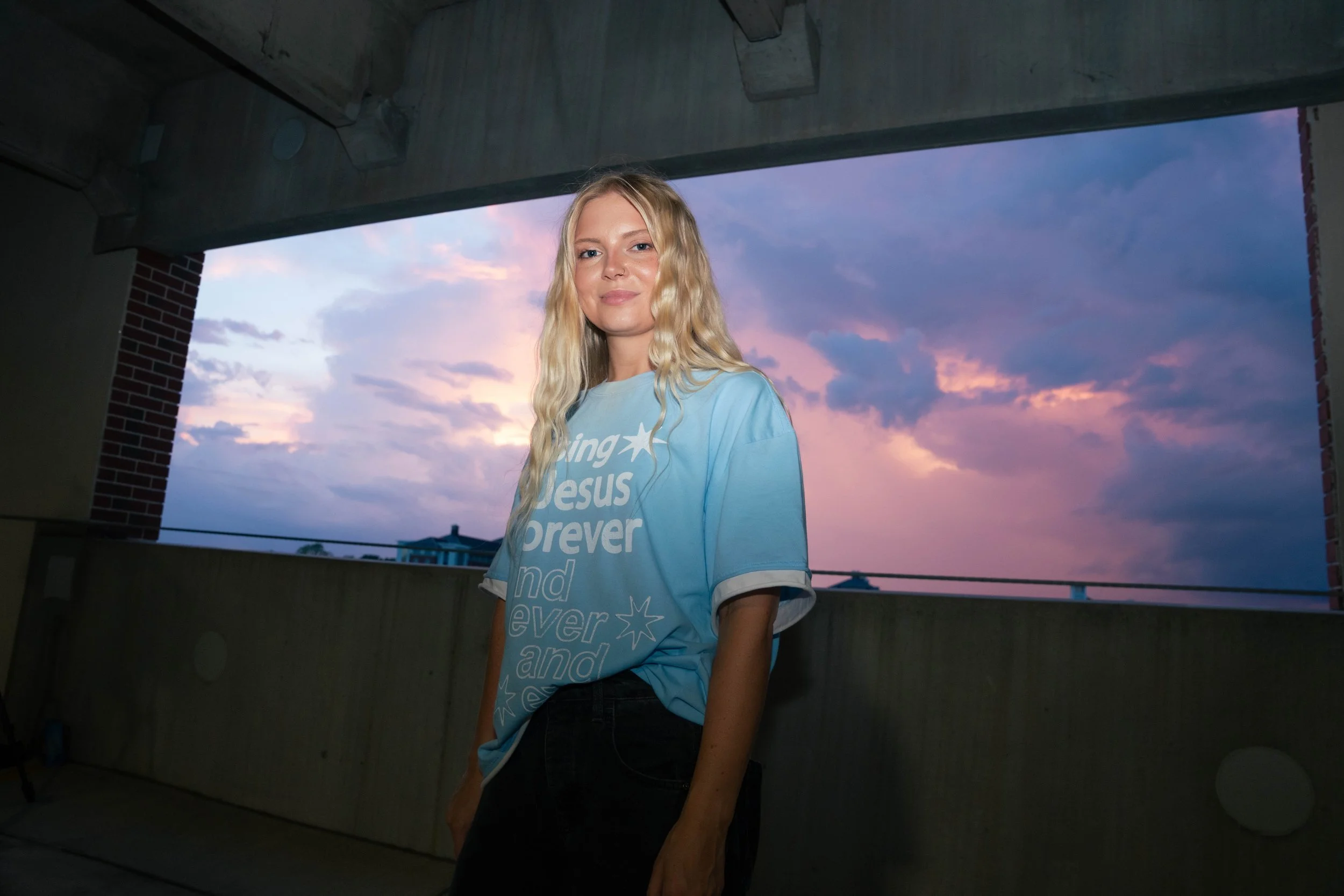 A young woman with blonde, wavy hair standing under a concrete structure with a scenic view of a colorful sunset sky in the background. She is wearing a light blue T-shirt with white text and a star, and black pants.