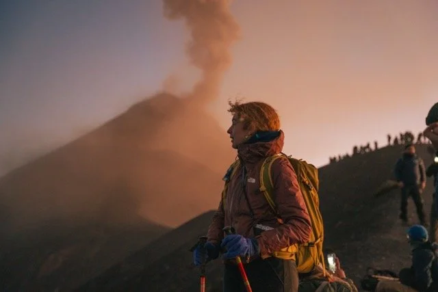 A woman with a backpack and walking poles standing on a mountain trail near erupting volcano with ash plume at sunset.