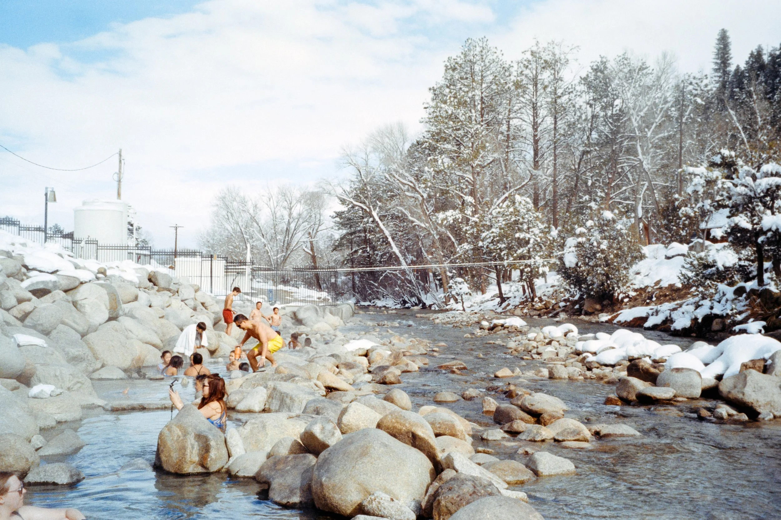 People swimming and relaxing in a rocky river during winter, with snow-covered trees and blue sky in the background.