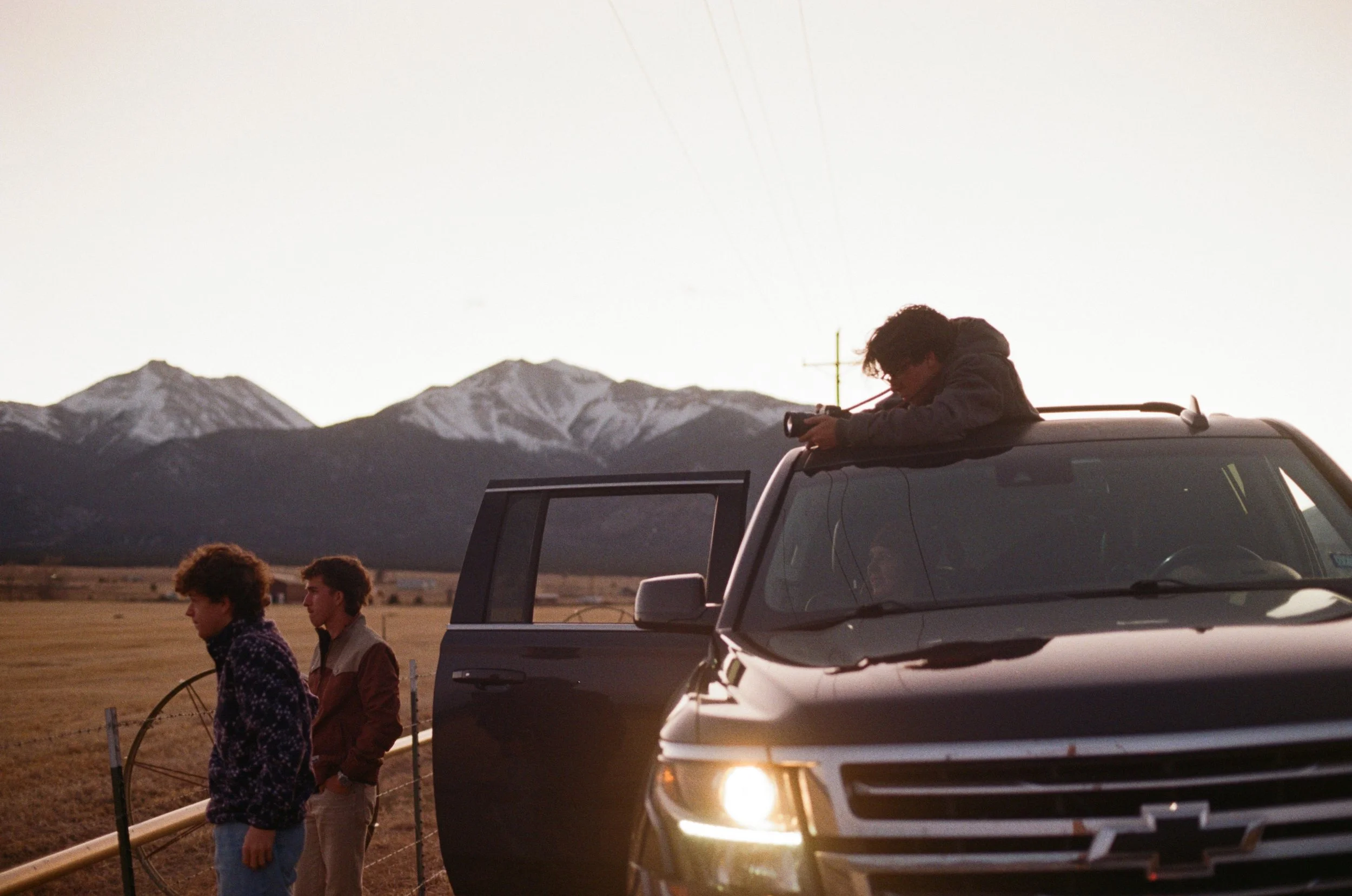 A person leaning out of a vehicle taking a photo with a camera, with mountains in the background, and a few other people walking nearby during sunset.