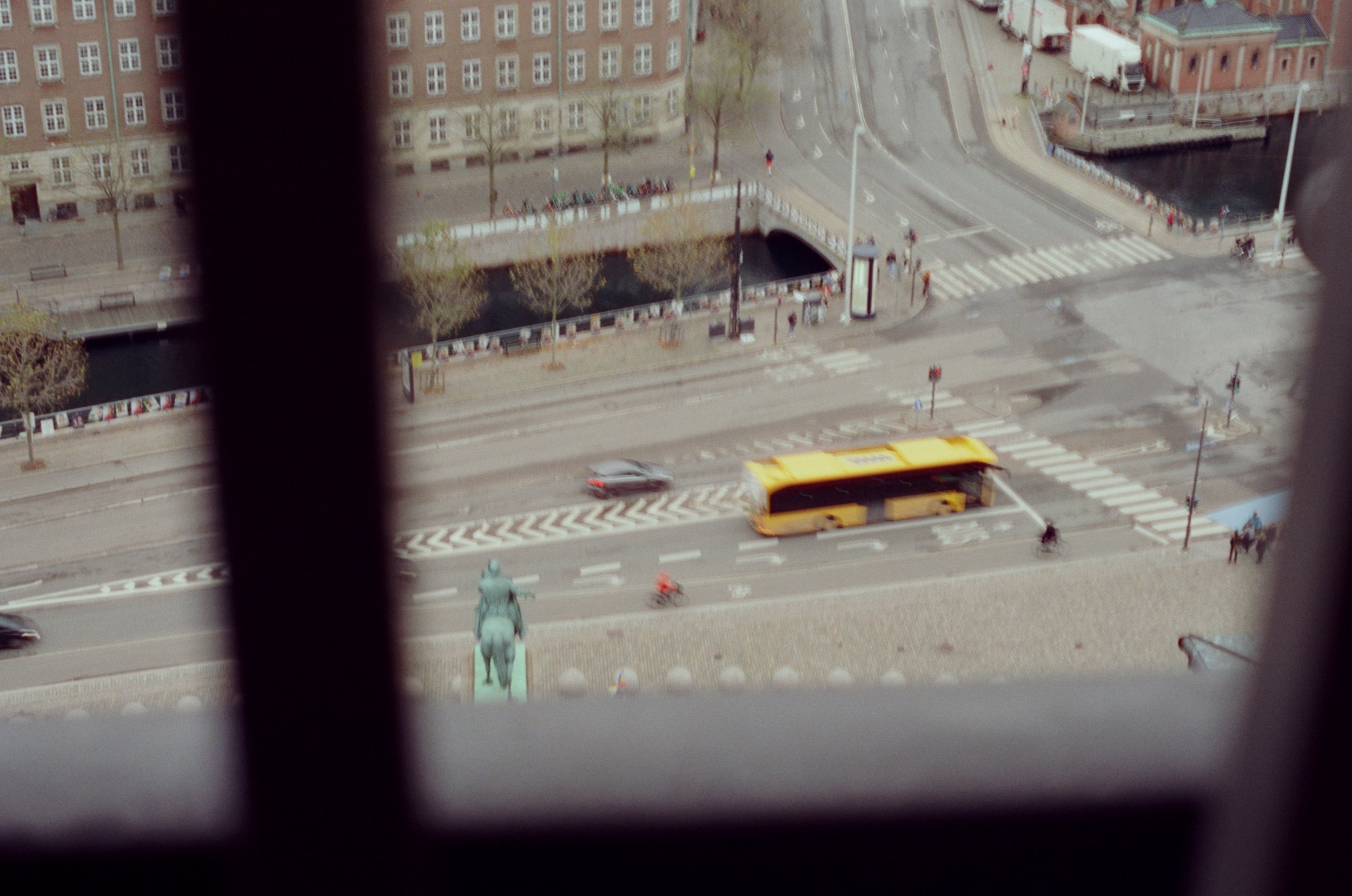 View from a building window shows a city street with a yellow bus, cars, cyclists, a monument, and a statue of a rider on a horse, with buildings and trees in the background.