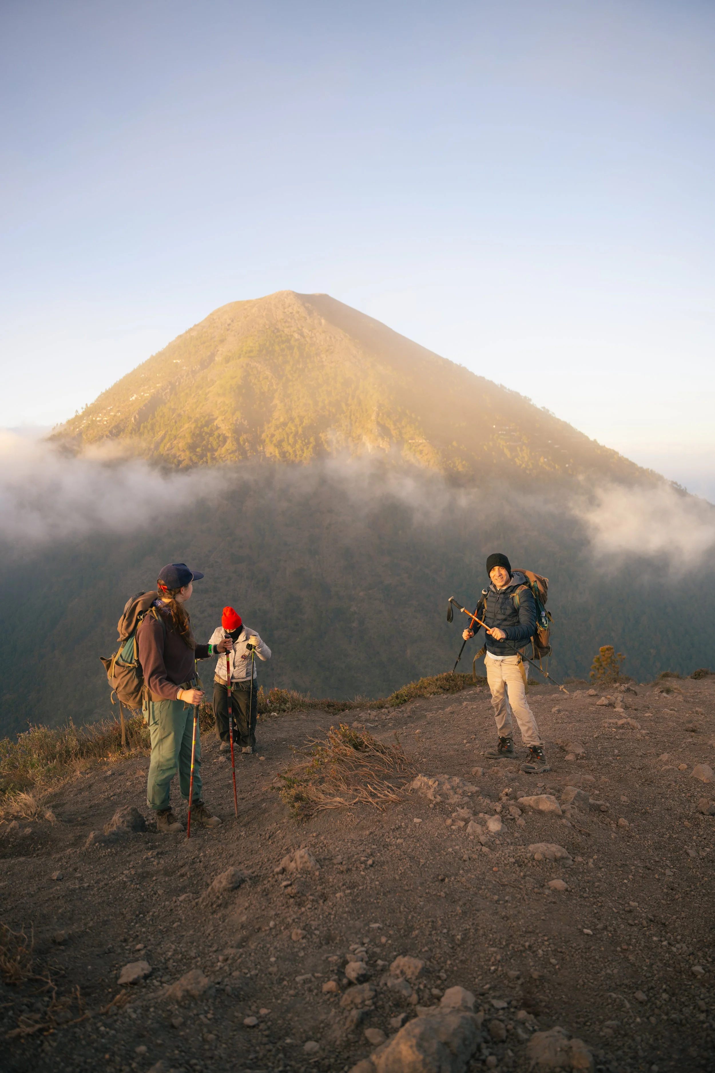 Group of hikers with backpacks and trekking poles on a mountain trail with a volcanic mountain in the background at sunrise or sunset.