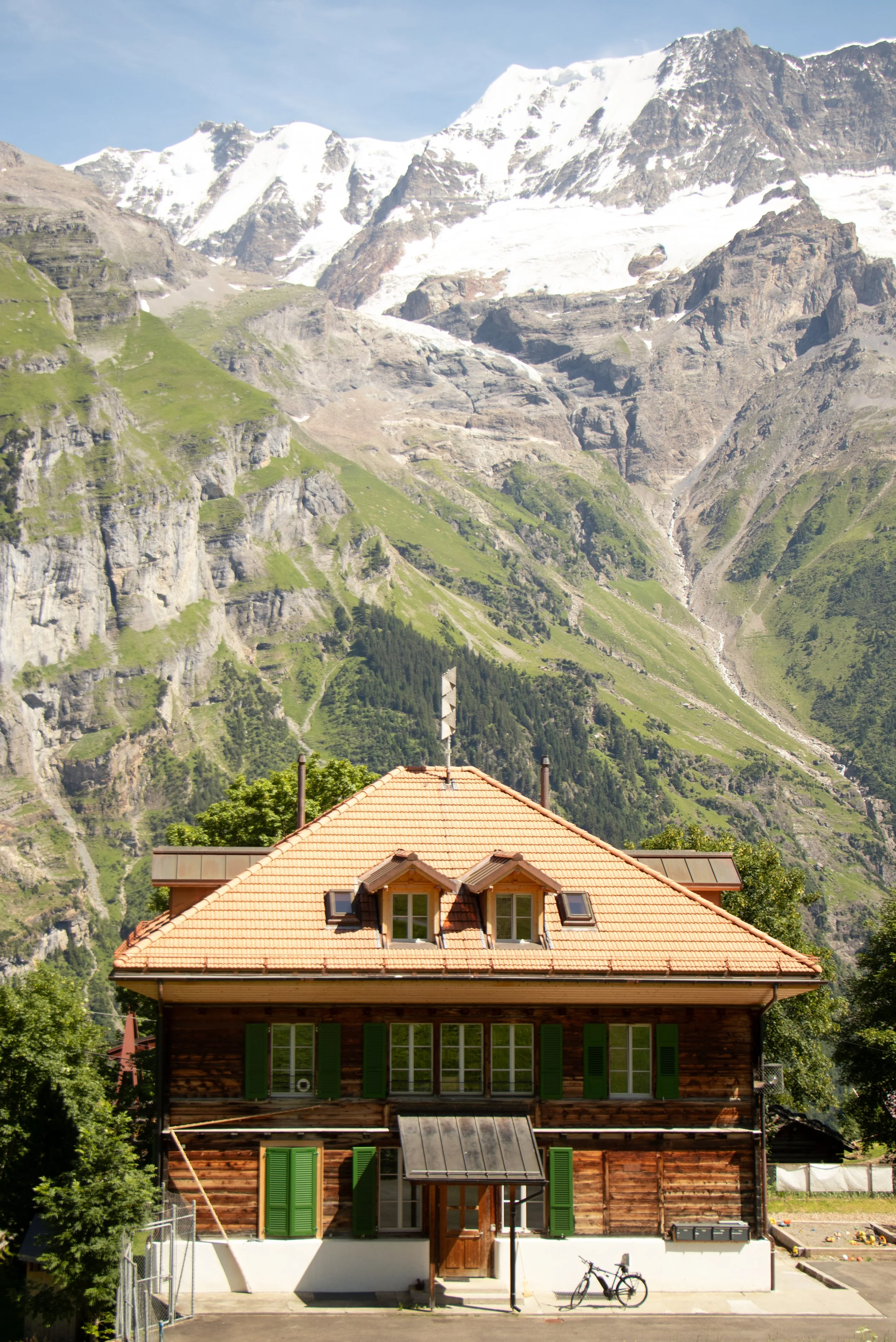 A house with a wooden exterior, green shutters, and a red tile roof, set in a mountainous landscape with snow-covered peaks in the background.