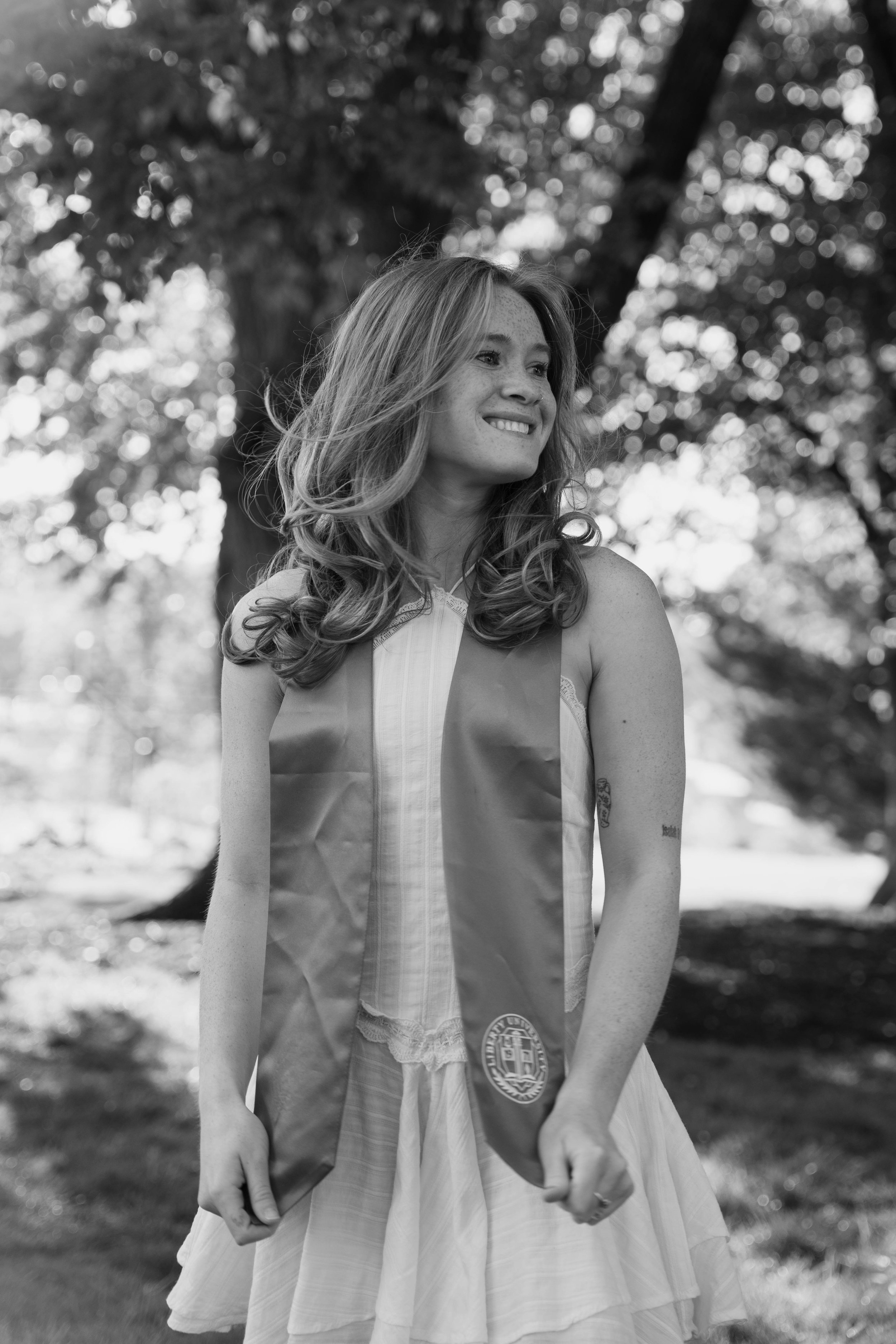 A young woman with long hair smiling outdoors, wearing a dress and graduation sash, with trees and blurred sunlight in the background.