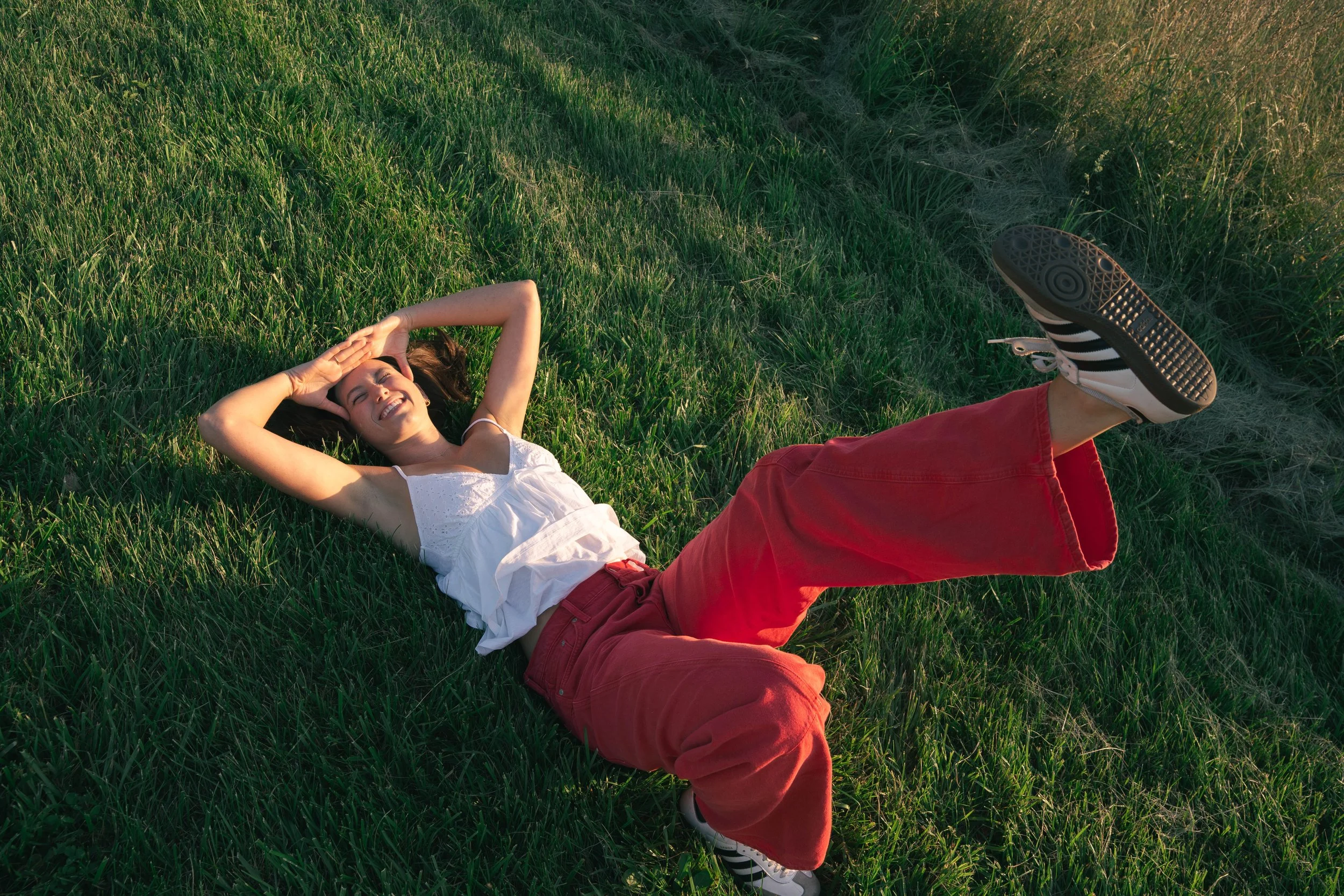 A young woman lying on green grass with a big smile, wearing a white tank top, red pants, and sneakers, enjoying the sunlight.
