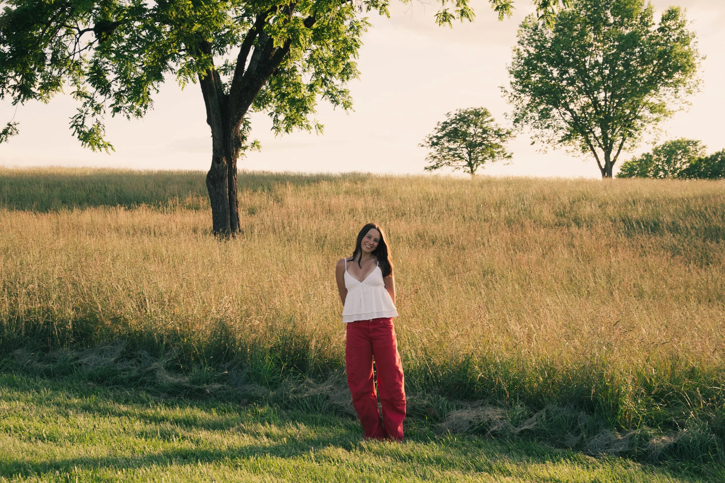 Woman standing in a grassy field with tall trees and a bright sky behind her, smiling.