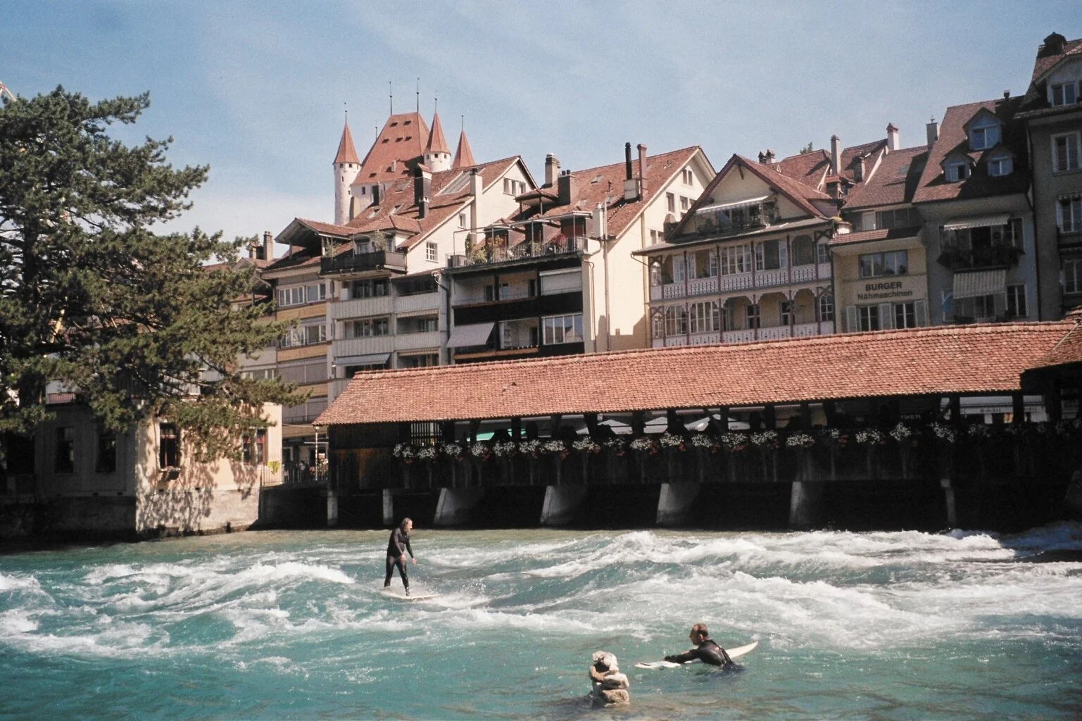 People surfing and paddling in a river with traditional European buildings along the riverbank in the background.