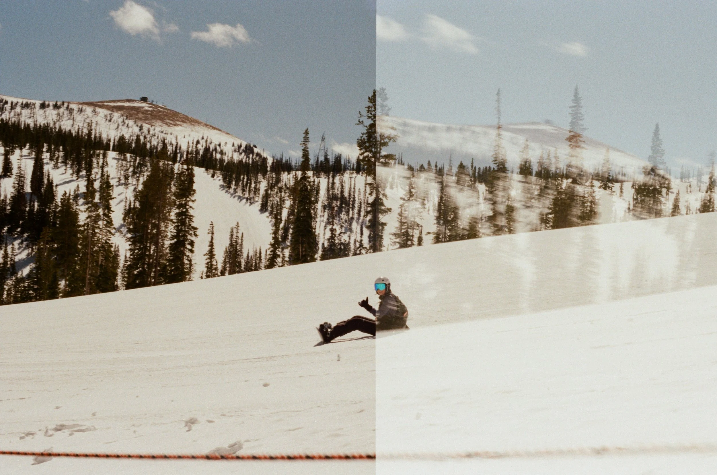 A person wearing ski gear and goggles sitting on snow with a snowy mountain and evergreen trees in the background.