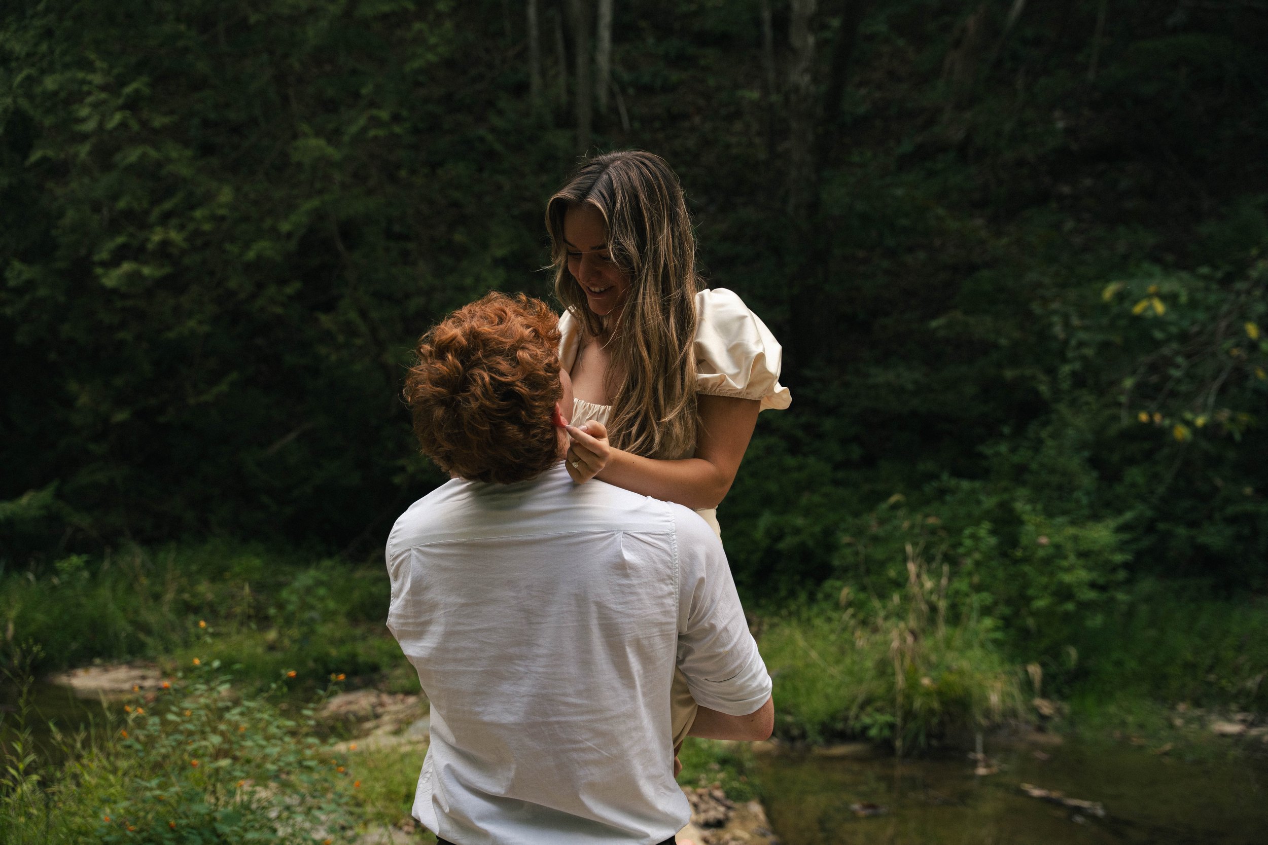 A couple is outdoors in a green, wooded area, with the man kneeling and the woman standing and smiling while holding his face, sharing a joyful moment.