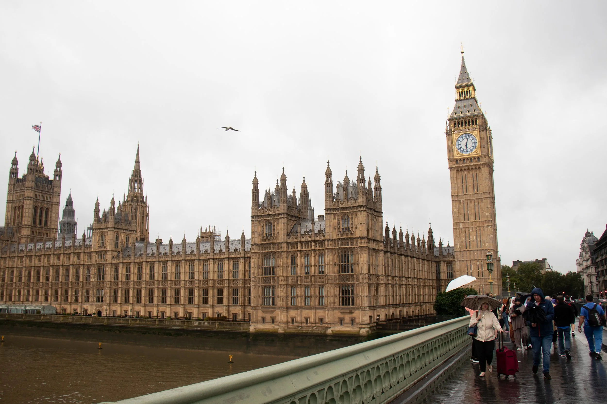 View of the UK Parliament building, known as the Palace of Westminster, with Big Ben clock tower, on a cloudy day. People are walking on a bridge with umbrellas, indicating rain.