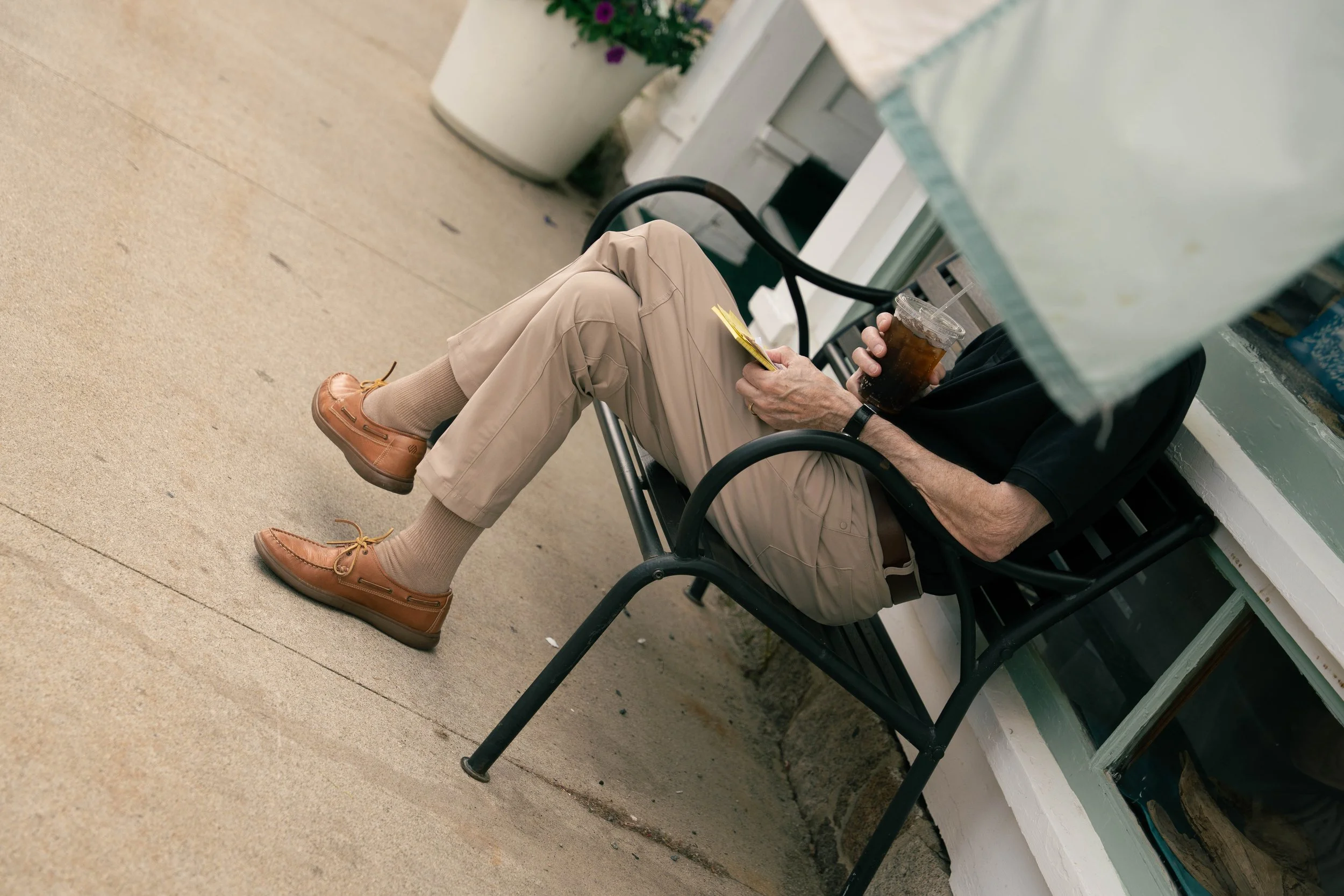 A person sitting on a black chair outdoors, wearing tan pants, brown shoes, and beige socks, holding a plastic cup of dark soda with ice, near a white building with potted plants.