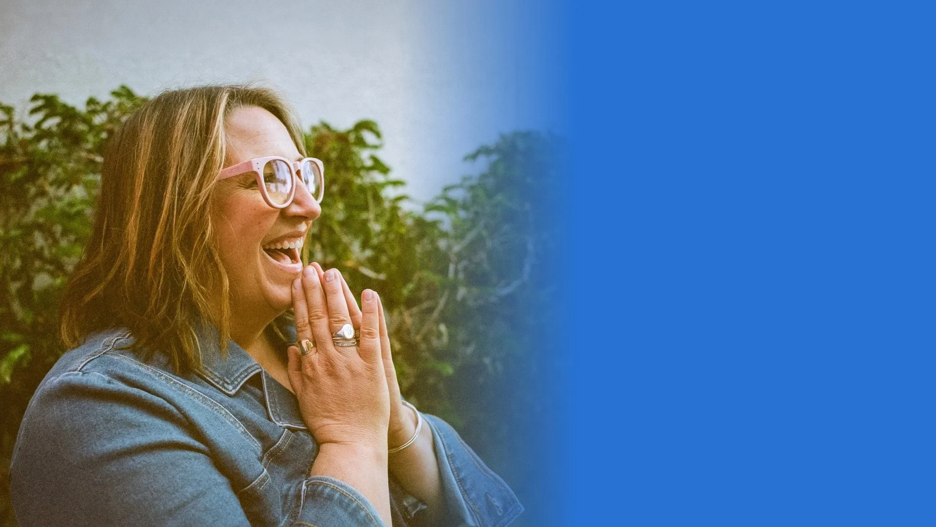 Molly Balint, wearing pink glasses and a denim shirt, smiling and laughing with her hands clasped near her face, outdoors with green foliage in the background.