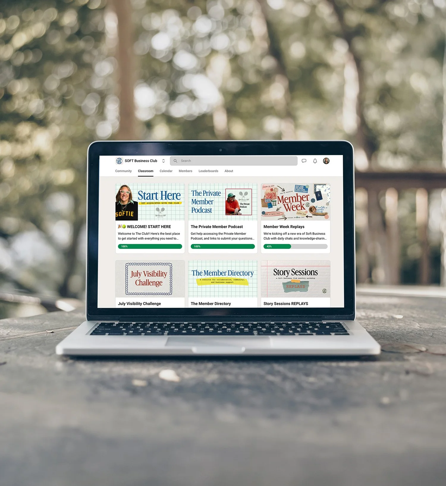 Open laptop displaying a digital classroom or community platform called SOFT Business Club, with various sections like 'Start Here', 'The Private Member Podcast', and 'Member Week Replays', set outdoors on a concrete surface with blurred trees in the background.