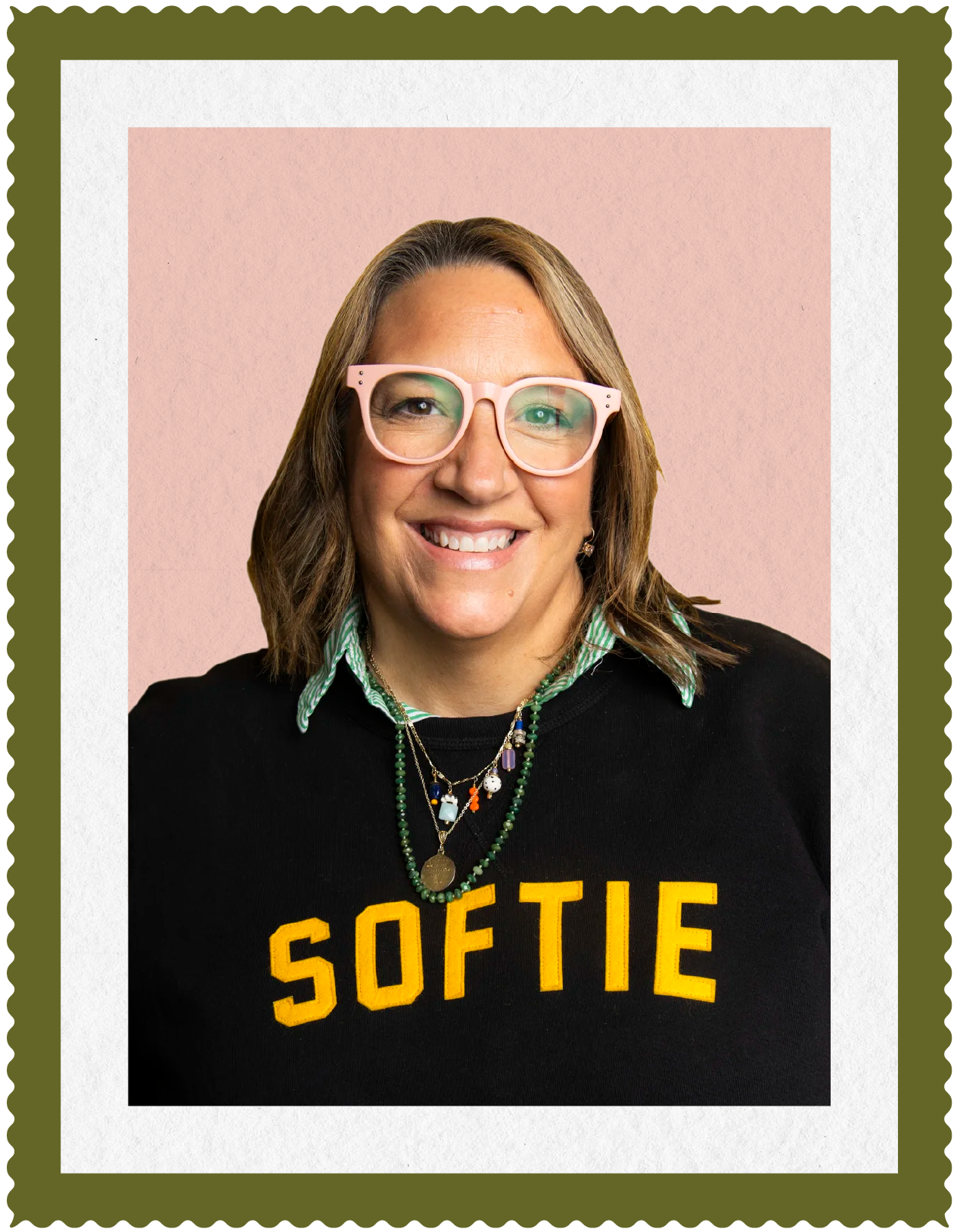 Molly Balint wearing pink glasses, a black sweatshirt with yellow text "SOFTIE," layered necklaces, and a striped collared shirt, set against a pink background with layered colored borders.