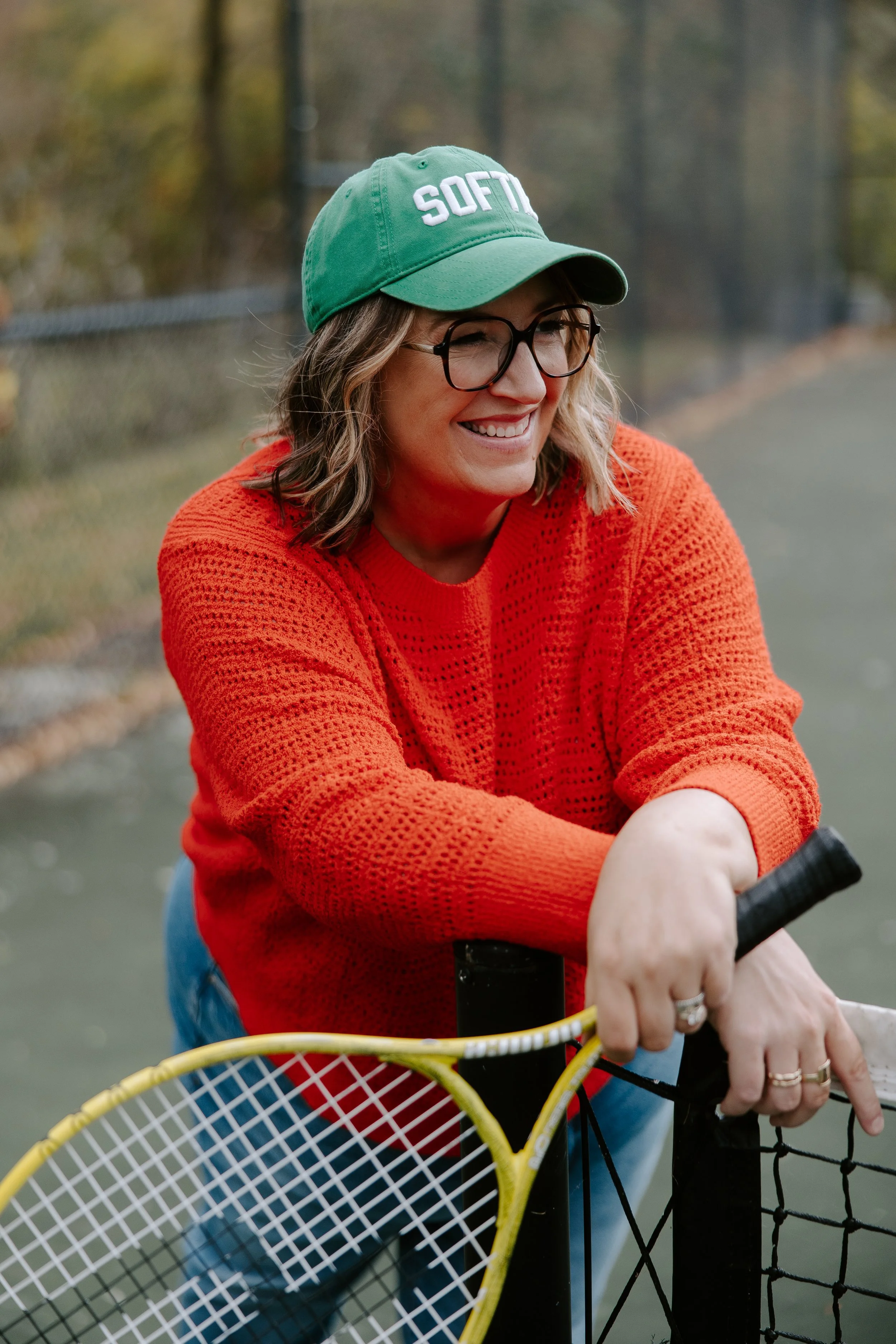 Molly Balint wearing glasses and a green cap with the word 'SOFTIE' on it, smiling while riding a tennis bike with a tennis racket in front of a fence and outdoor tennis court, autumn trees in the background.