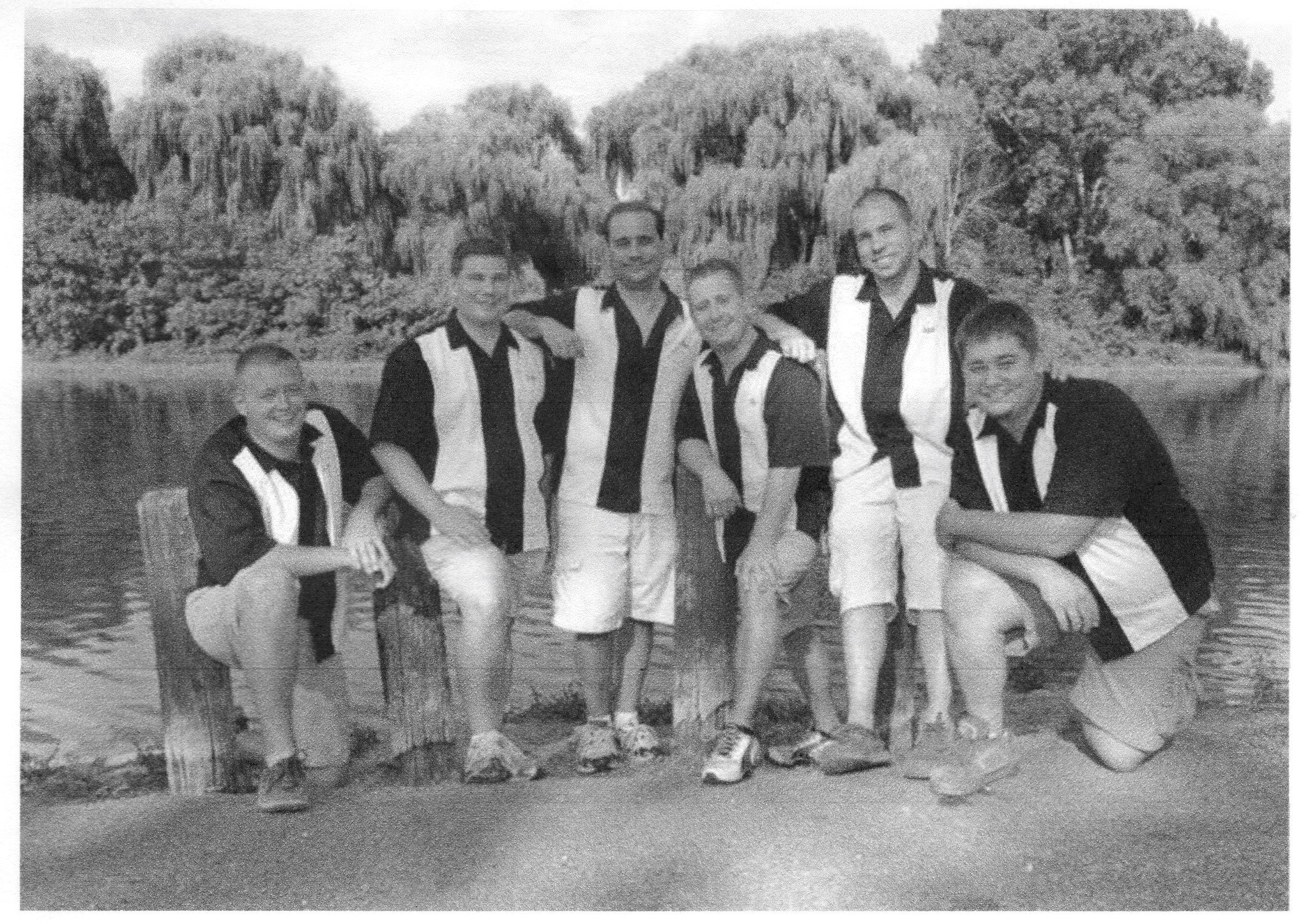Black and white photo of six young men standing outdoors in front of trees, some holding their hands on the other’s shoulders, all dressed casually.