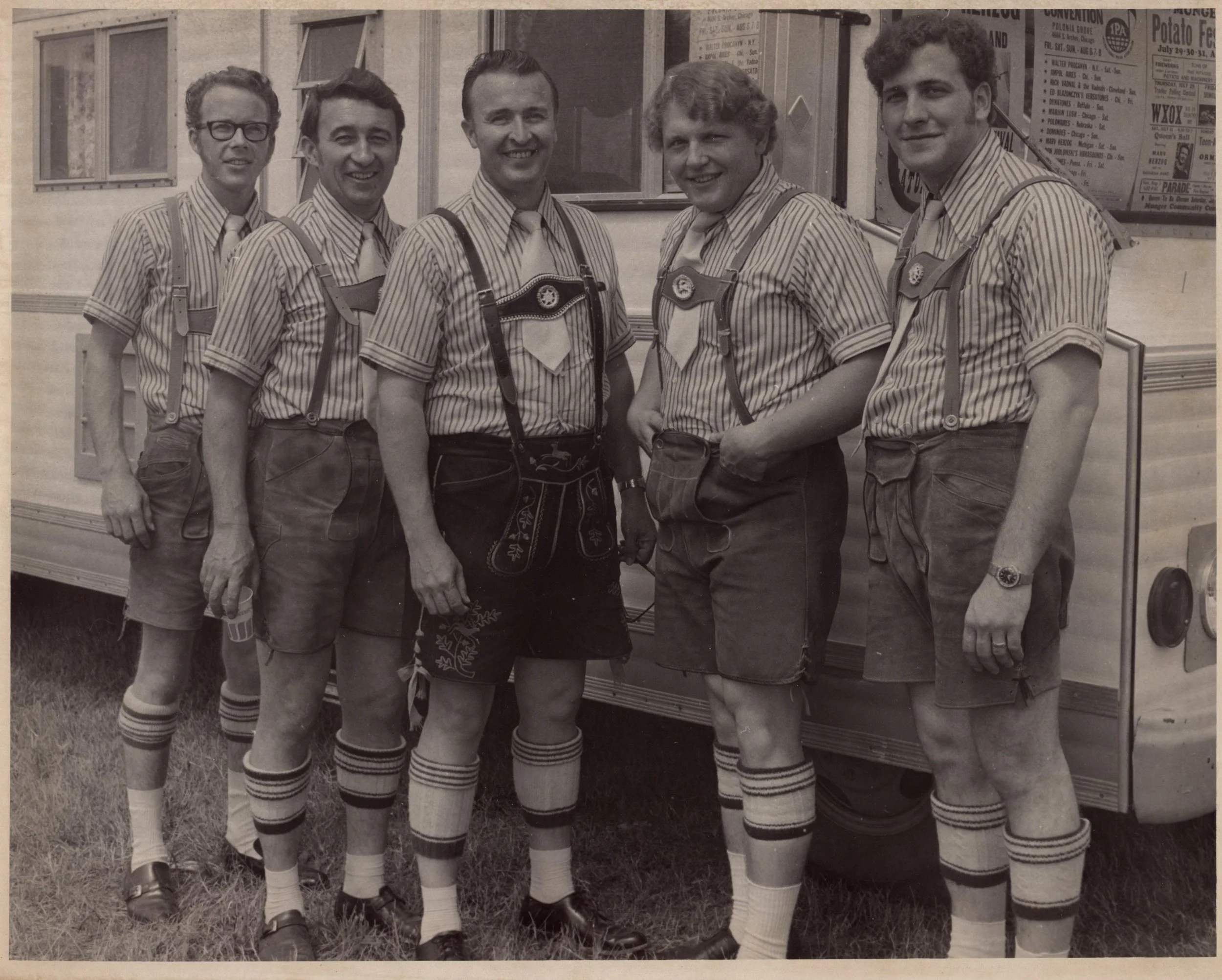 Black and white photo of six men in retro sports uniforms consisting of striped shirts, suspenders, shorts, long socks, and sneakers, standing outdoors in front of a trailer with posters on its exterior.