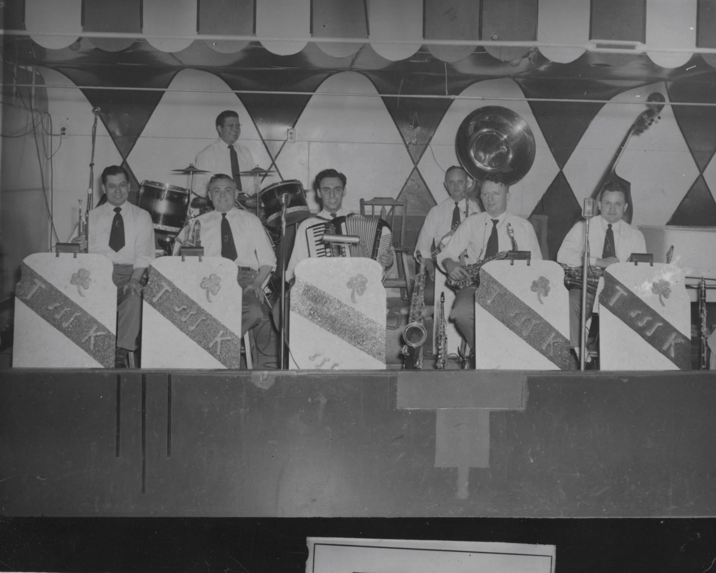 Black and white photo of a hand-painted mural of a band of six musicians with musical instruments, in front of a background with triangular patterns and draped fabric with shamrock designs.