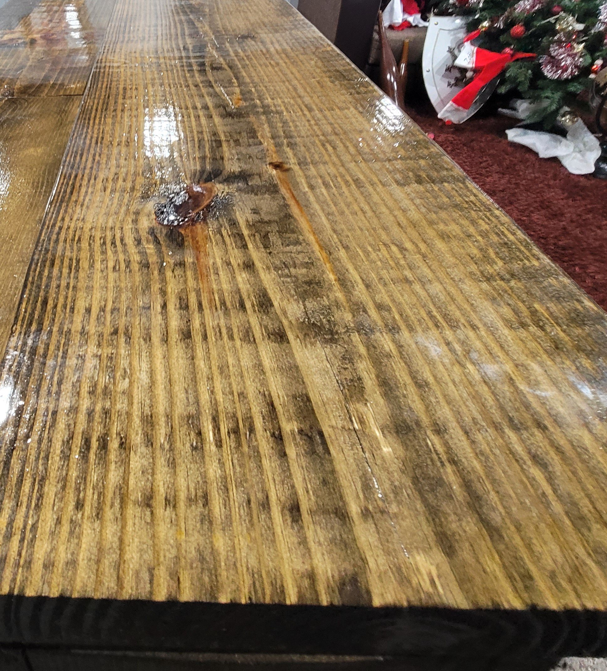 Close-up view of a wet, polished wooden table with visible wood grain, and a small shiny leaf placed on it. In the background, part of a decorated Christmas tree and some wrapped packages are visible.