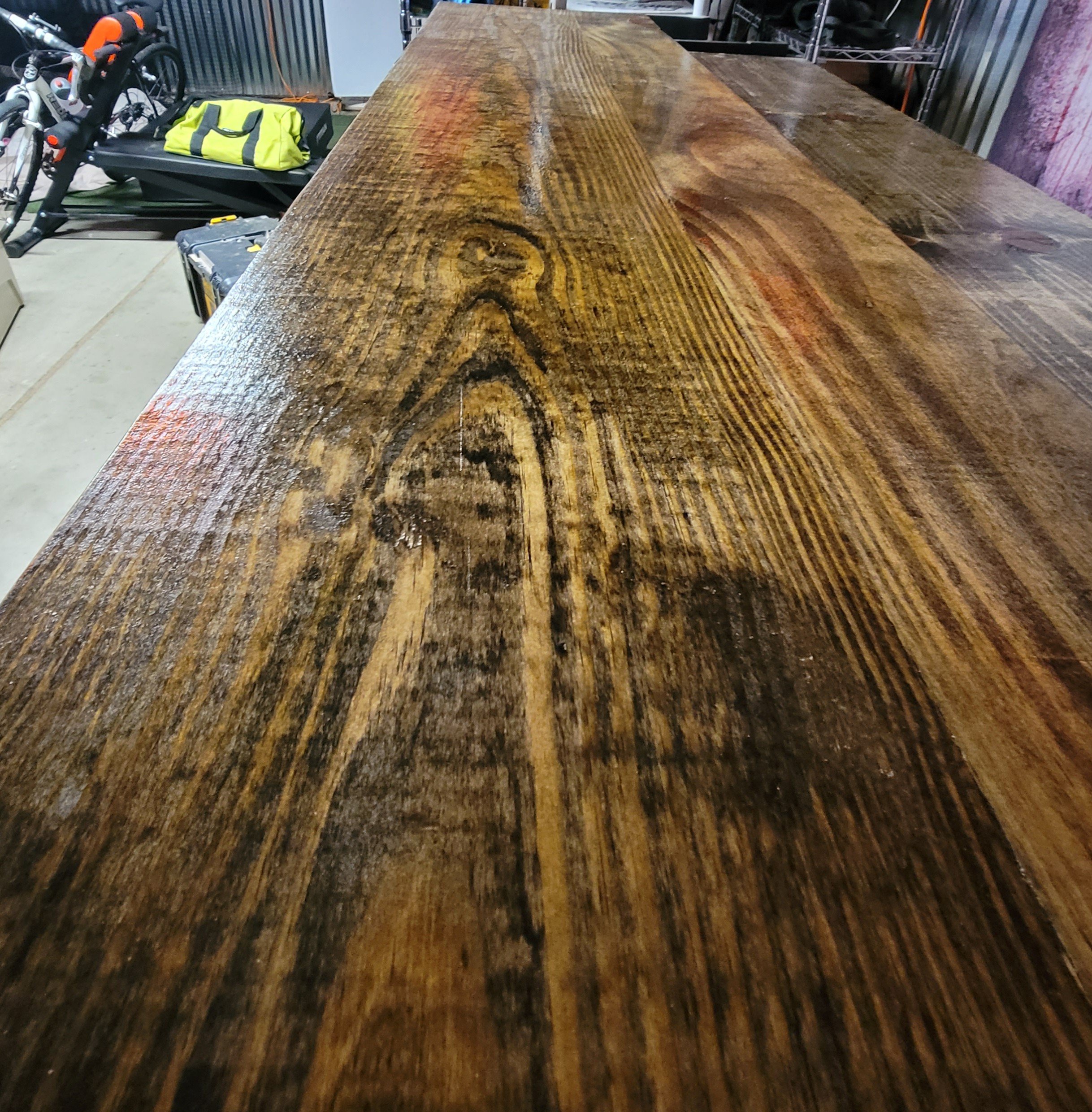 A long, polished wooden table in a storage or workshop area, with bicycles, a yellow bag, and tools visible in the background.