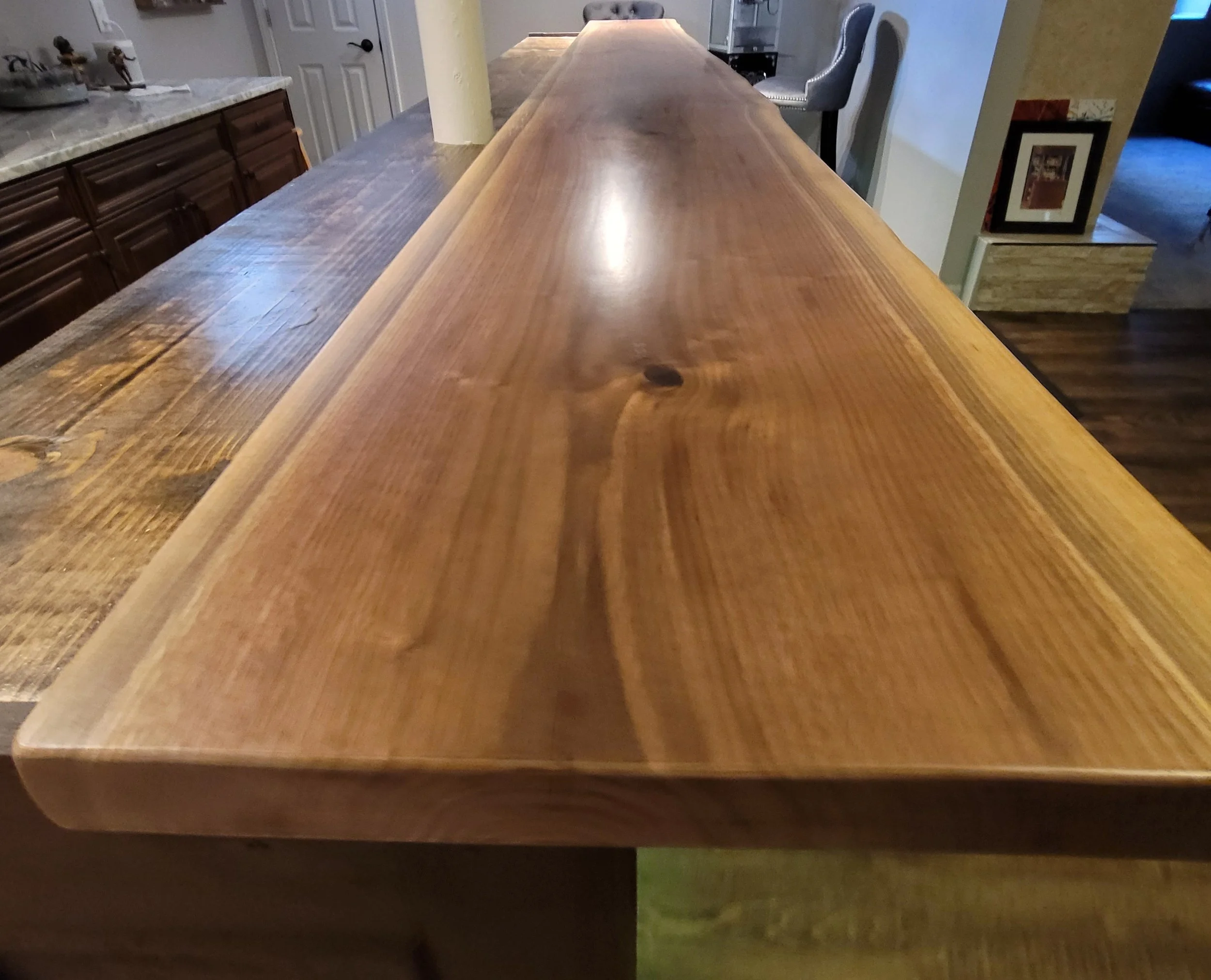 A close-up image of a wooden countertop or table with a smooth, glossy surface, with visible wood grain and knots. In the background, part of a kitchen area with cabinets and a decorative chair are visible.