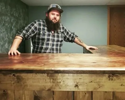 Man with beard and plaid shirt leaning on a wooden bar counter.