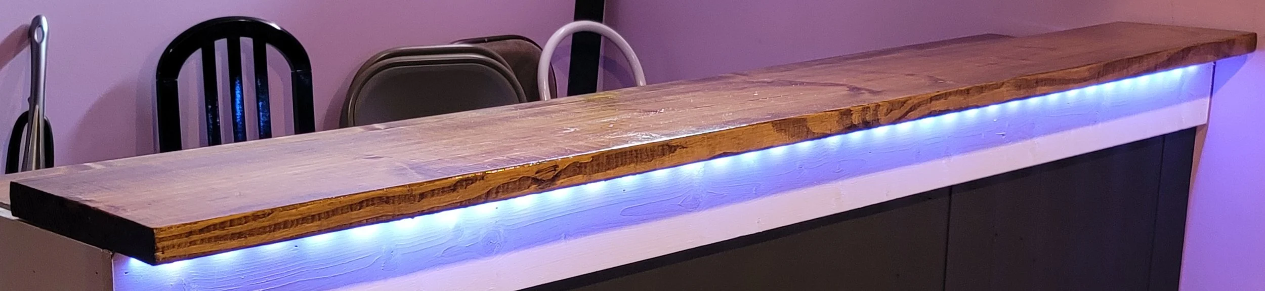 A wooden bar top with blue LED lighting underneath, against a pink wall with several chairs behind it.