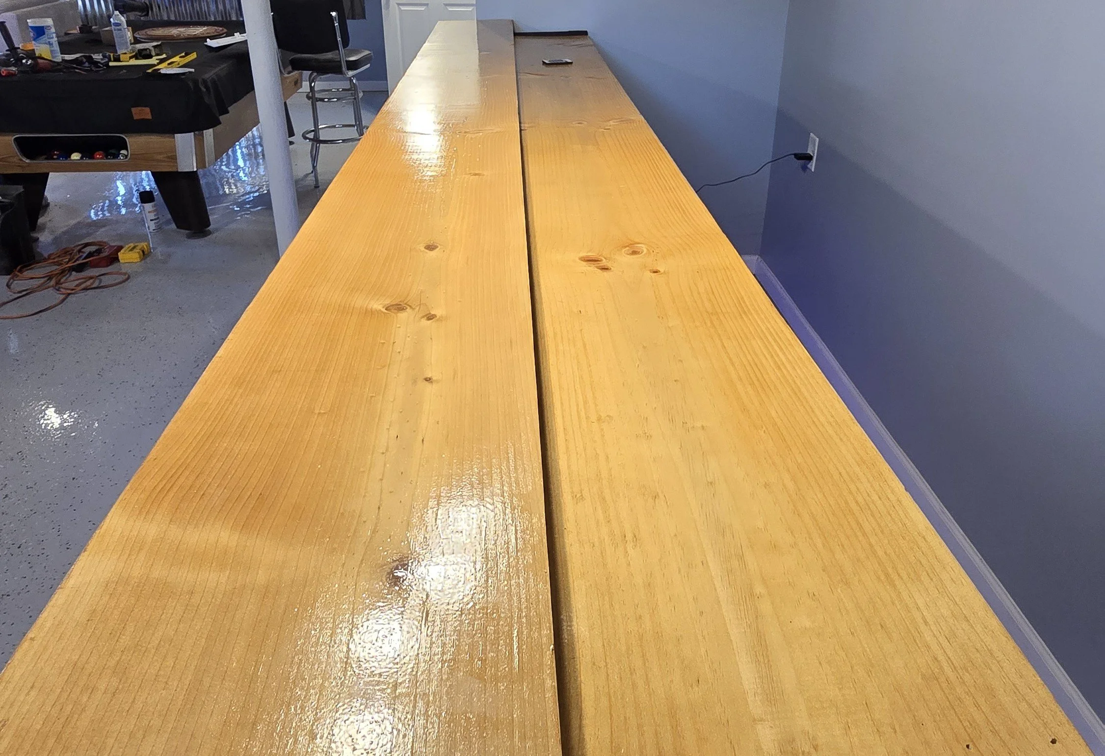 Long wooden table with a glossy finish in a room with blue-gray walls, power outlet on the wall, and some objects including a cell phone on the table.