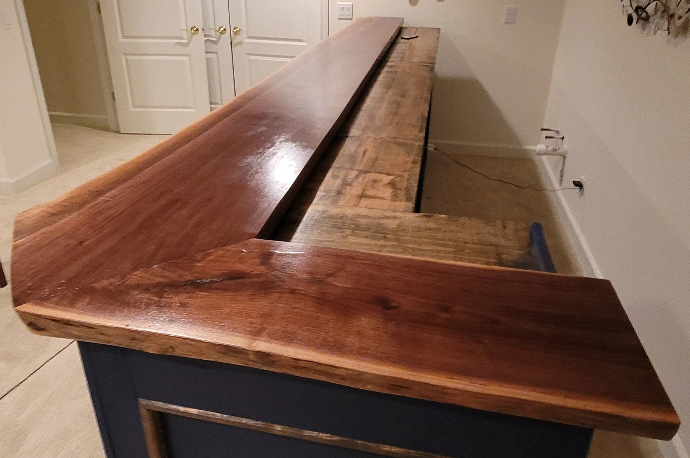 A wooden table with a natural finish surfaces and a smooth, polished top with visible wood grain. The table is situated in a room with plain light-colored walls, next to a corner with a wall-mounted coat rack and electrical outlets visible.