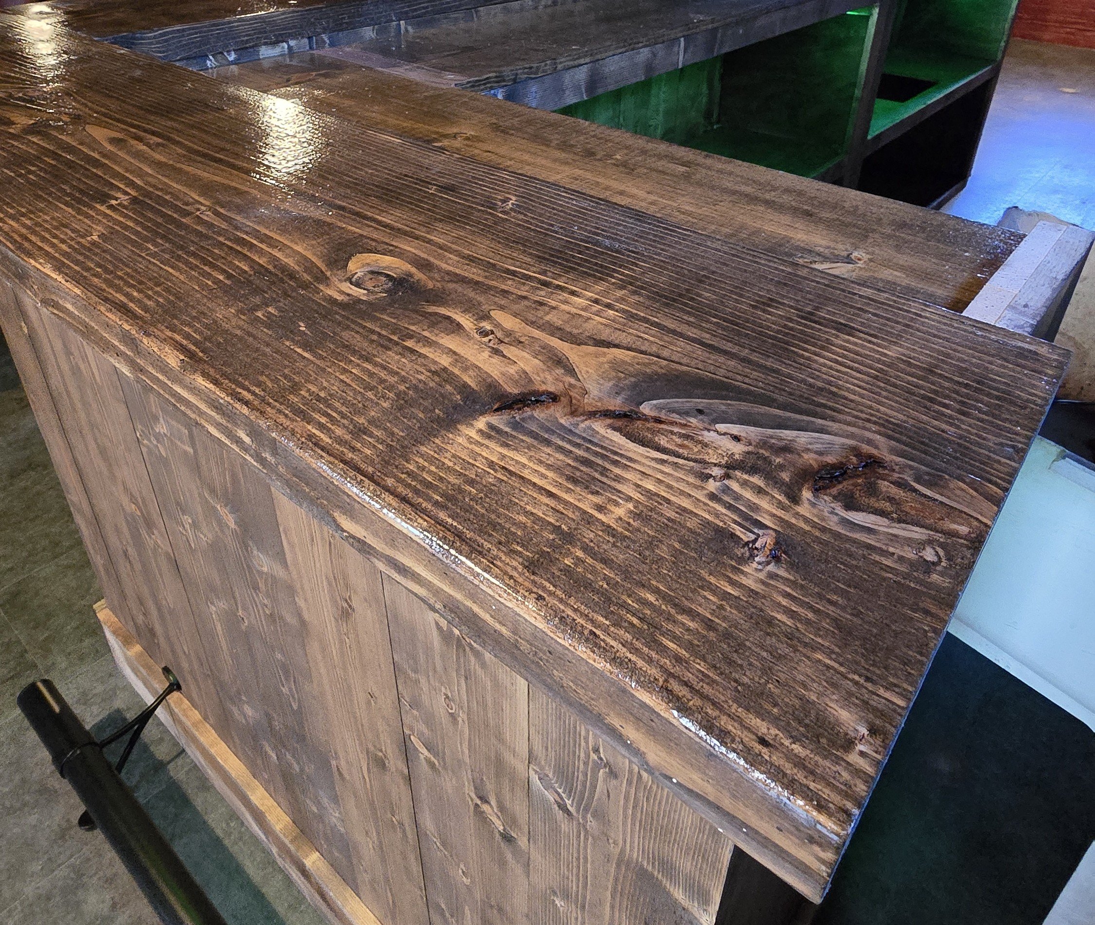 This image shows a close-up of a wooden table with a shiny, polished surface. The table is part of a furniture piece with a natural wood grain pattern and knots visible. In the background, there is another piece of furniture with a green and black design, and part of a dark brown surface is also visible.