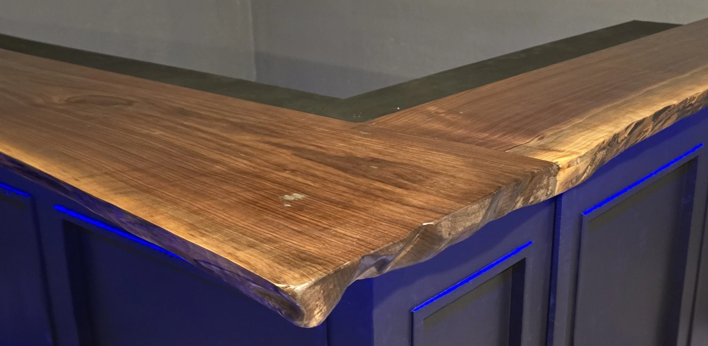 Close-up of a kitchen countertop made of wood with a natural edge, illuminated by under-cabinet lighting, with blue cabinet doors visible beneath.