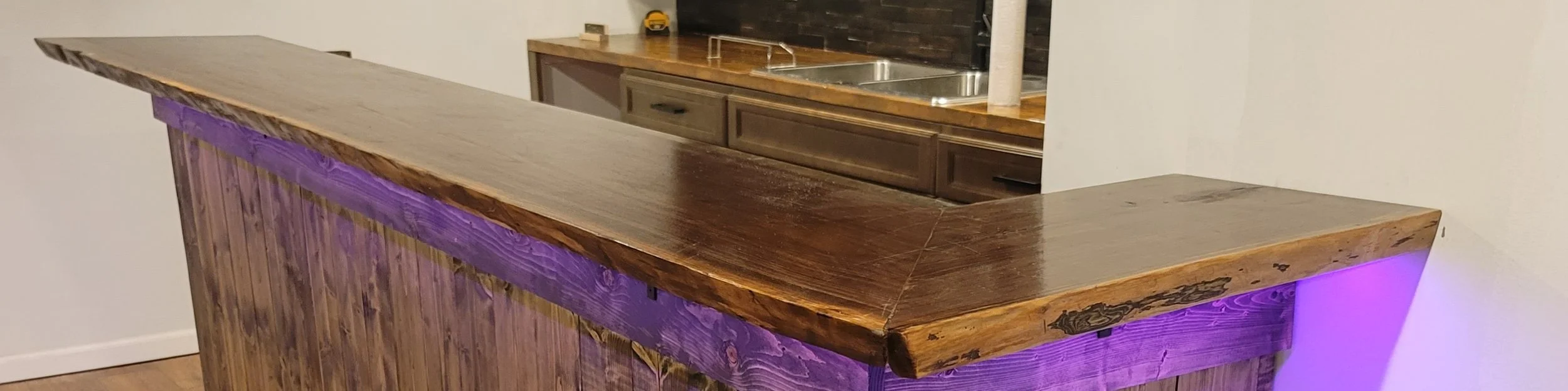 Wooden bar counter with purple lighting underneath, in front of a kitchen area with a sink and cabinets.