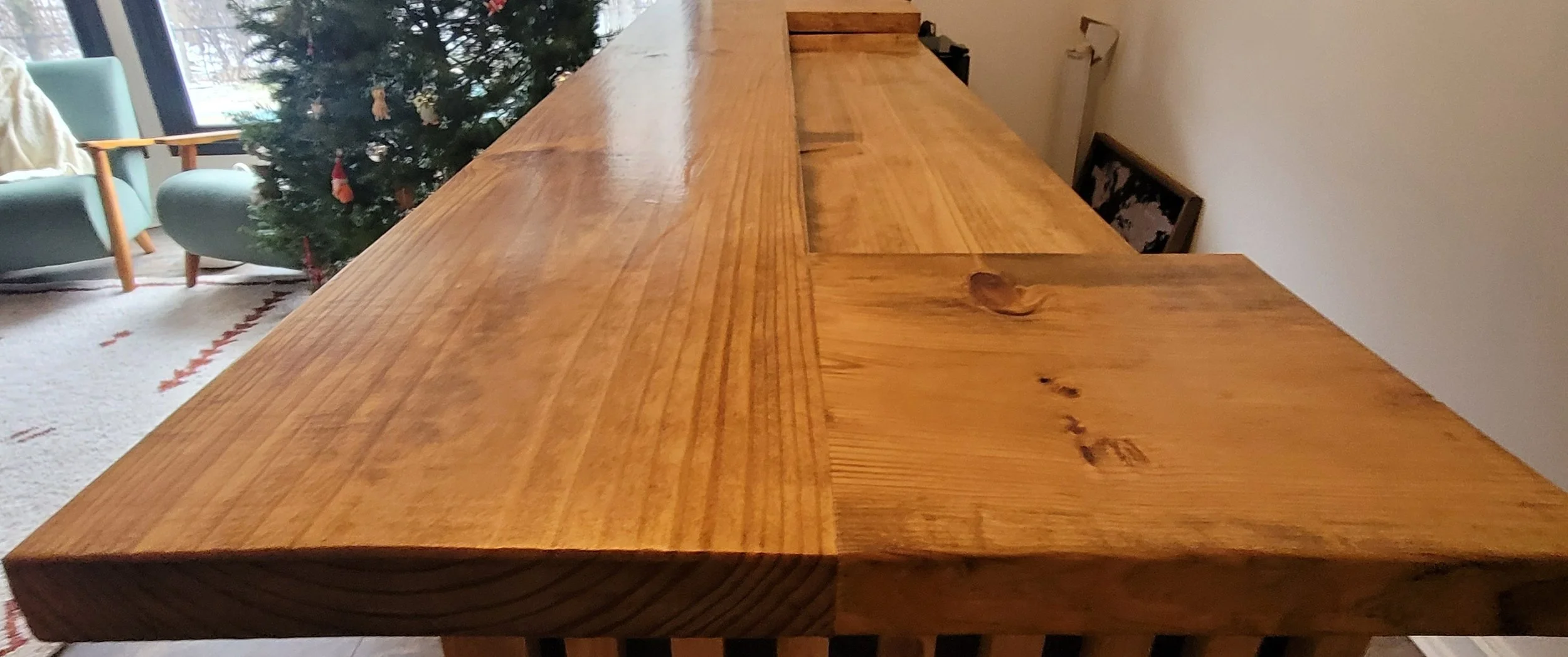 Close-up of a large wooden dining table with visible wood grain and knots, situated in a room with natural light coming from a window or sliding door in the background.