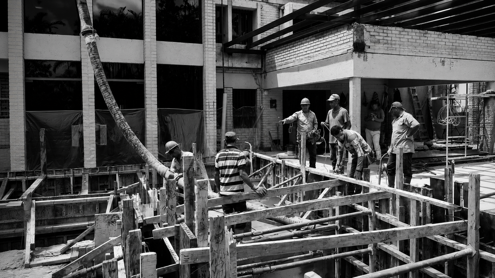 Construction workers on site working on a building foundation, with wooden formwork and concrete pipes visible.