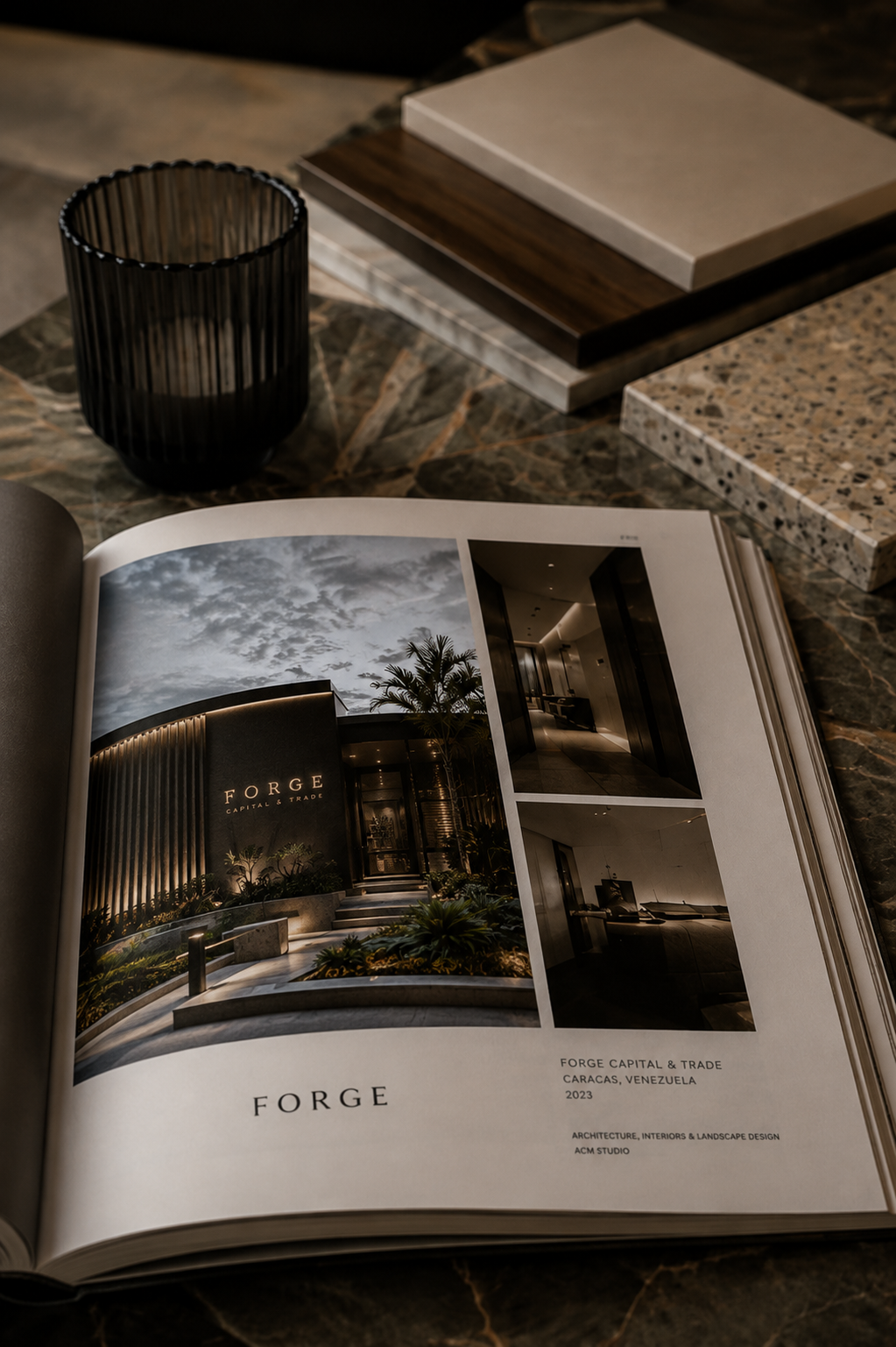 Open architecture and design book featuring images of a modern building facade with the text "FORGE" and location "Caracas, Venezuela" on a marble table. Surrounding items include a black cup, sample tile and material swatches.