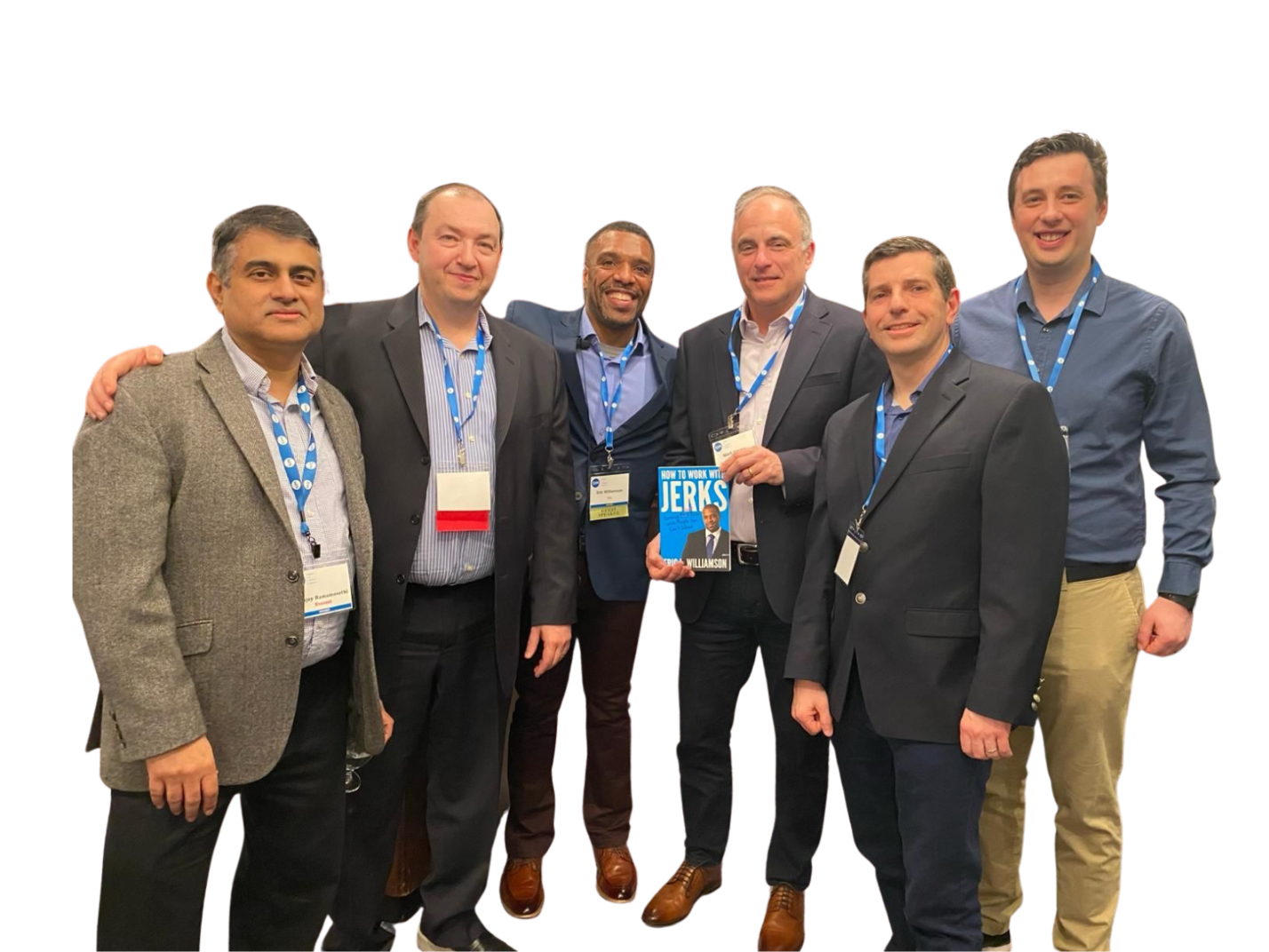Group of six men in formal attire posing for a photo at a conference, with one man holding a book titled 'How to Work with Jerks' by Michael B. Williamson.