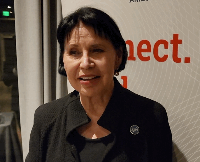 A woman with short black hair wearing a black blazer and a round pin, standing in front of a beige background with red and black text.