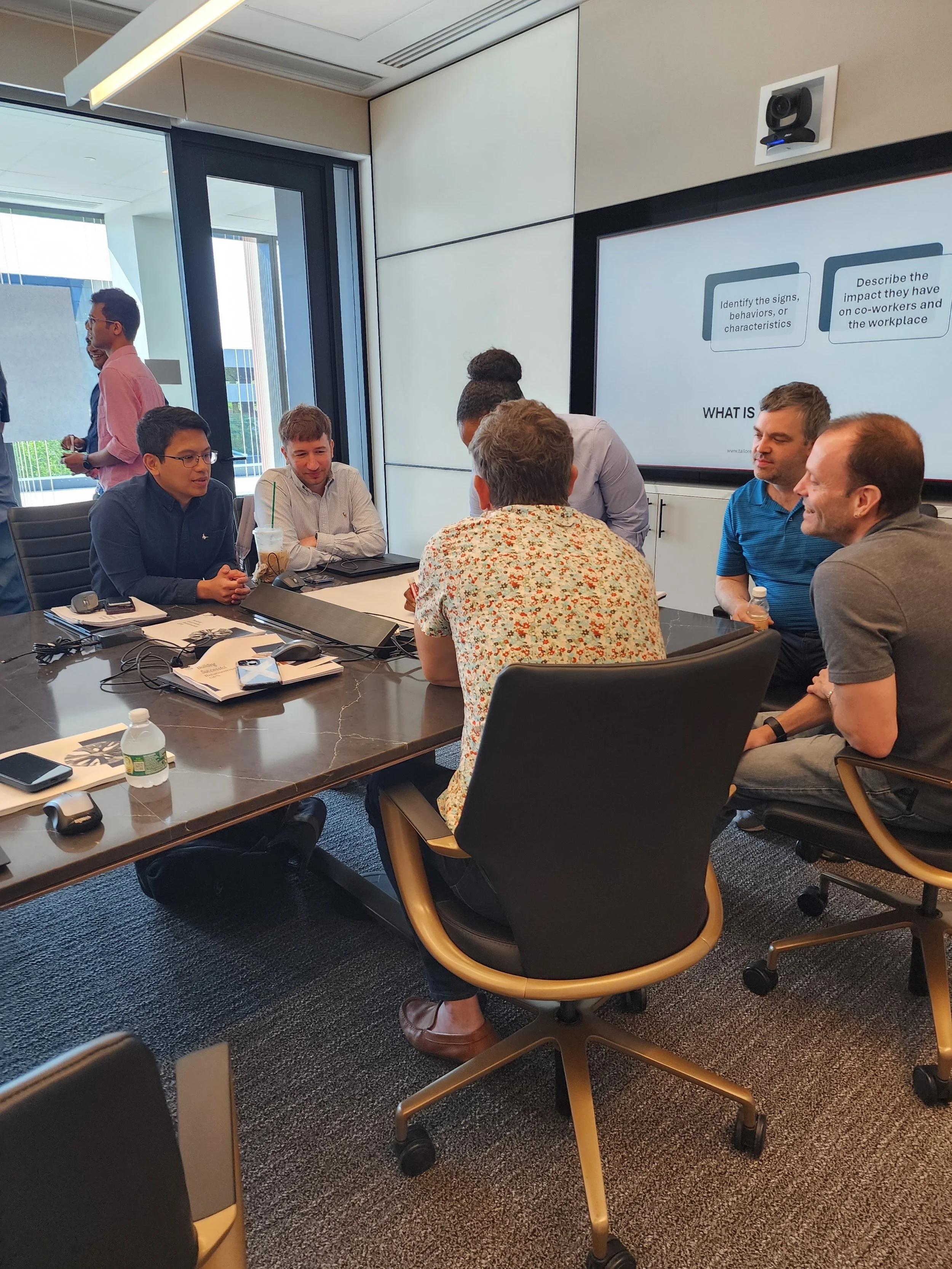 Six people are sitting around a conference table in a modern office, engaged in discussion. There are notebooks, a phone, and drinks on the table, with a large screen displaying presentation slides behind them.
