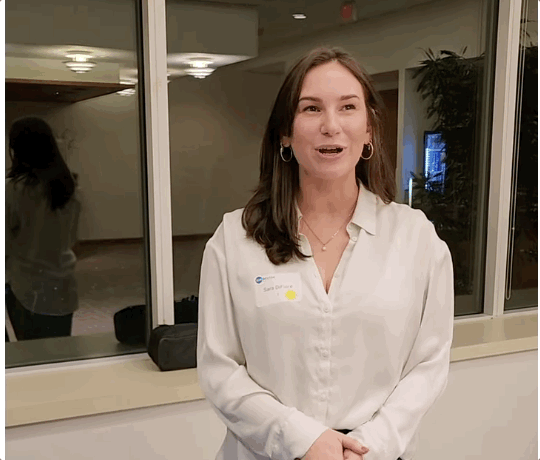 A young woman with brown hair, wearing a white blouse and earrings, standing indoors near a large window, talking to someone.