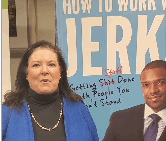 A woman with dark hair wearing a black turtleneck, blue blazer, and pearl necklace standing in front of a blue poster with partially visible text and a man in a suit.