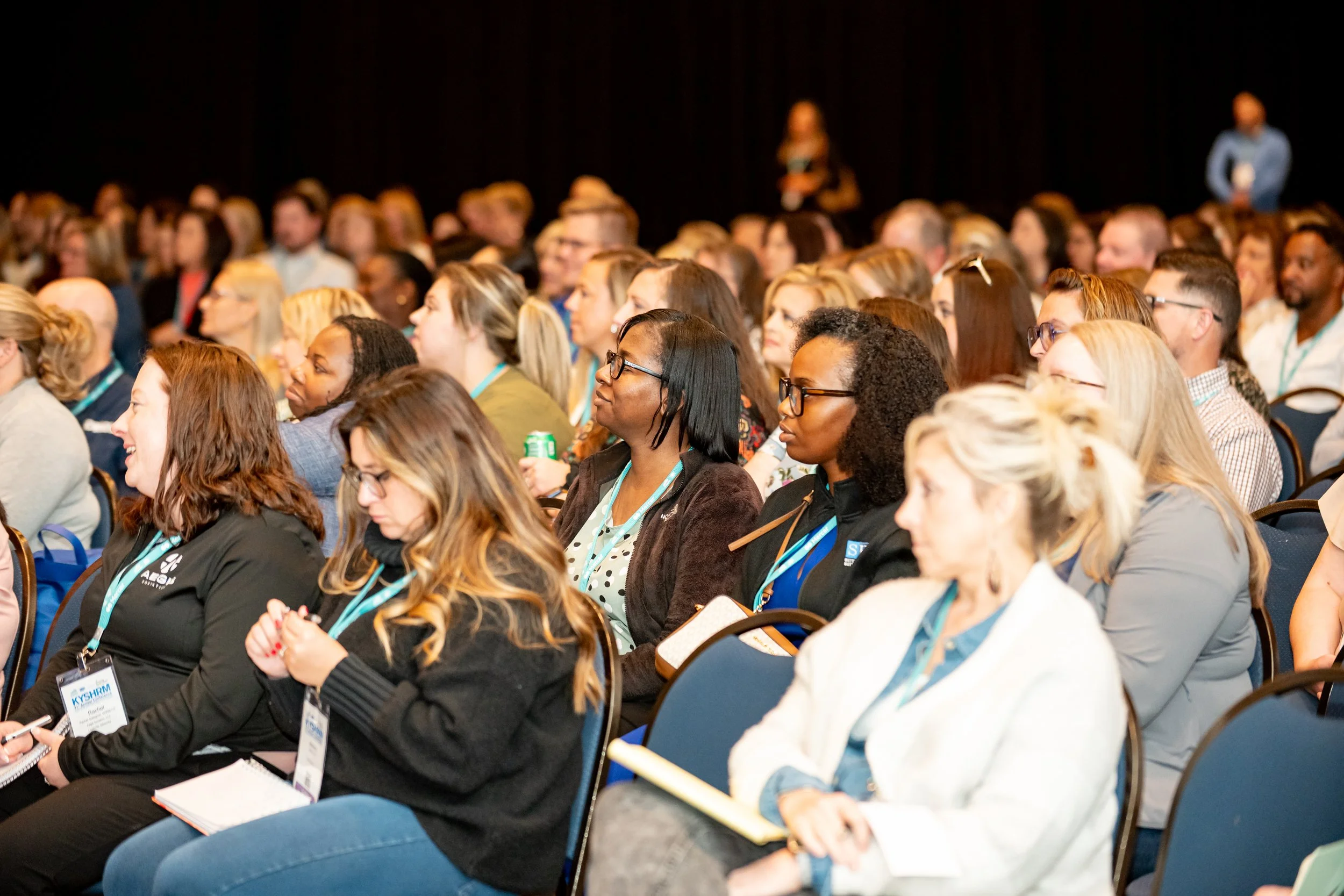 A large audience of diverse individuals attending a conference or seminar, sitting in rows with some taking notes and wearing conference badges.