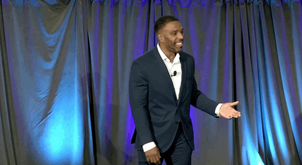 Man in suit giving a presentation with a microphone in front of a blue-lit curtain.