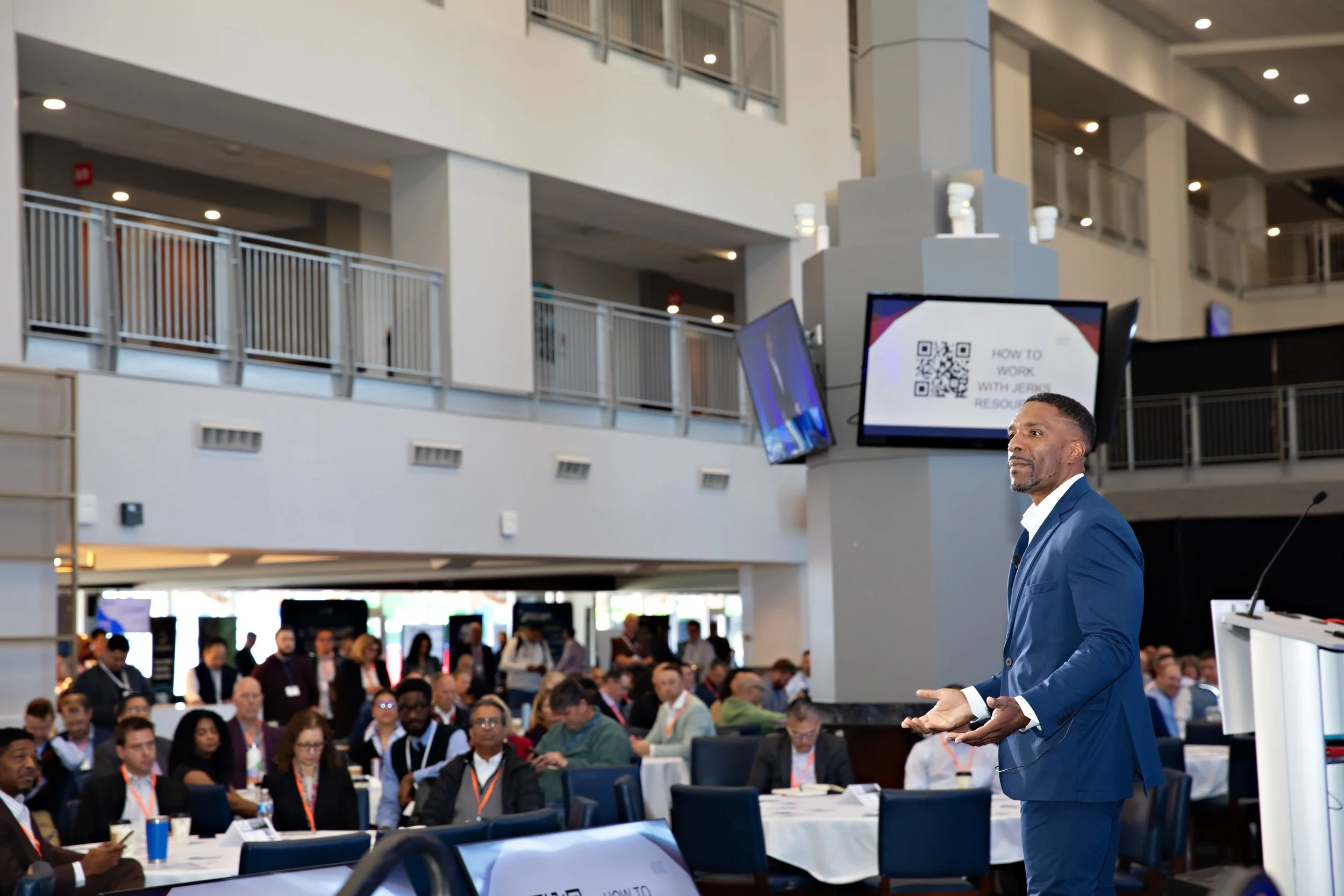 A man in a blue suit giving a presentation in front of a large audience at a conference or seminar.