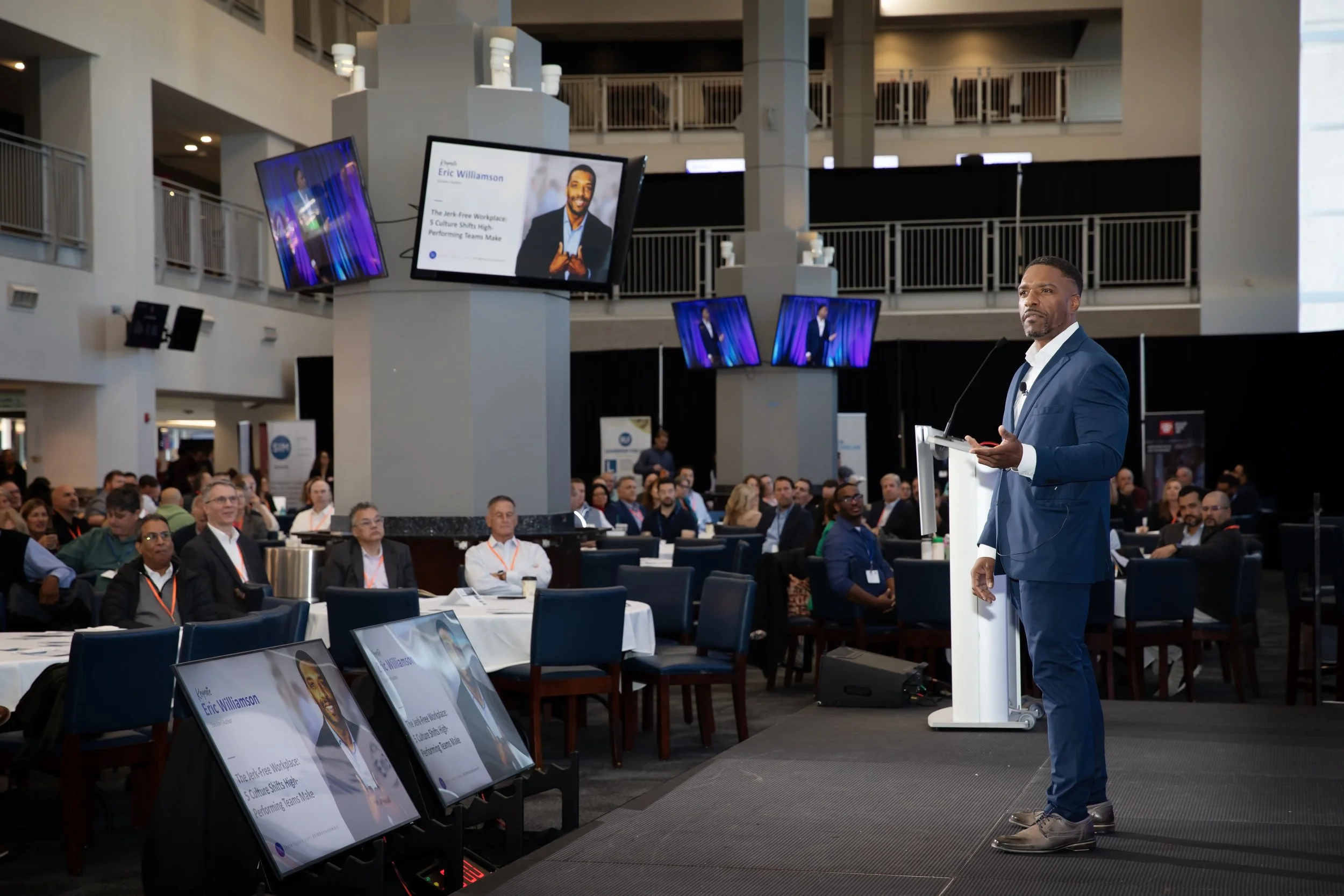 A speaker in a blue suit presenting at a conference with an audience seated at tables. Multiple screens display his image and presentation slides in a large indoor venue.