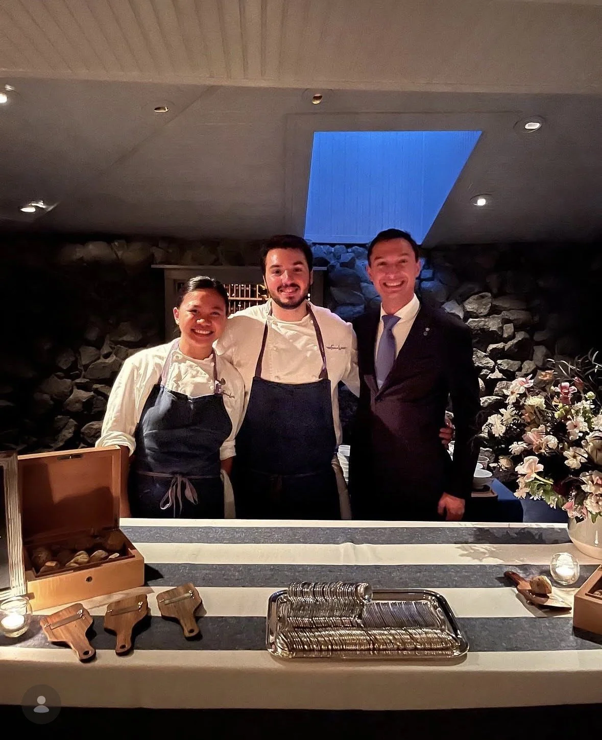 Three people standing together behind a table in a restaurant, smiling at the camera. Two of them are wearing chef uniforms with aprons, and one man is in a suit. The table has wooden utensils, a silver tray, and a floral arrangement.